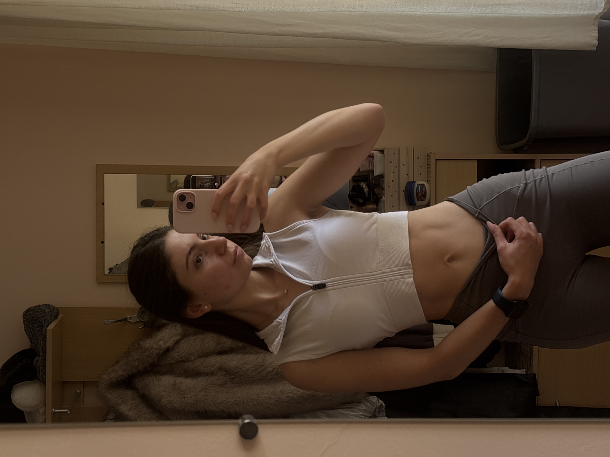A woman is standing in front of a mirror, taking a selfie in what appears to be a bedroom. She is wearing a white, sleeveless zip-up crop top and grey leggings. Her hair is dark and pulled back, and she has freckles on her face. She is wearing a small necklace and a smartwatch on her left wrist. The background shows a closet with clothing hanging, shelves with various items, and a trash can. A sheer curtain is visible on the right side of the image. The lighting suggests it is daytime. The location is not specified to be Varna, Bulgaria.