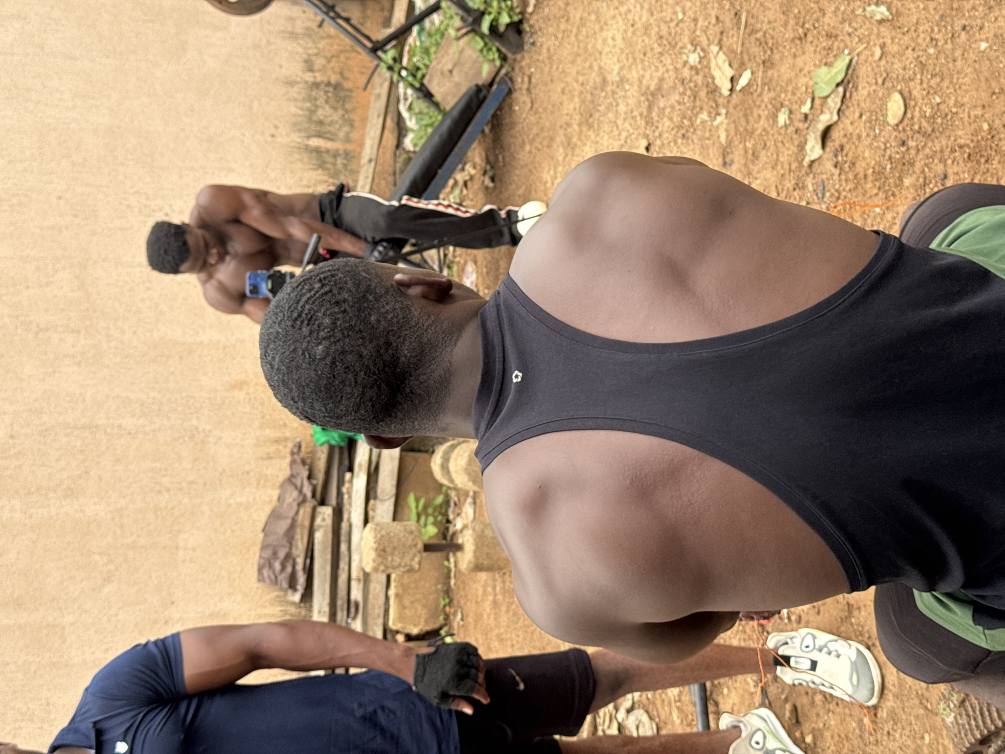In Akwanga, Nigeria, three Black men are engaged in a workout session outdoors. The foreground features a man from the rear, wearing a black tank top and dark shorts with a green accent. He is seated, facing away from the camera, with his muscular back and shoulders prominently displayed. To his left, another man stands, viewed from the side and rear, dressed in a dark blue polo shirt and black shorts. He wears black fingerless gloves. In the background, a third man, shirtless and with defined musculature, is captured mid-action, holding a smartphone and appearing to record or take a photo. He is wearing black pants with white and red stripes and white sneakers. The environment is rustic, with a sandy ground covered in fallen leaves and some sparse vegetation. In the background, there are stacked wooden planks, makeshift weights, and what appears to be a basic weightlifting bench. The wall behind them is a textured, light brown surface. The lighting suggests an overcast or shaded daytime condition.