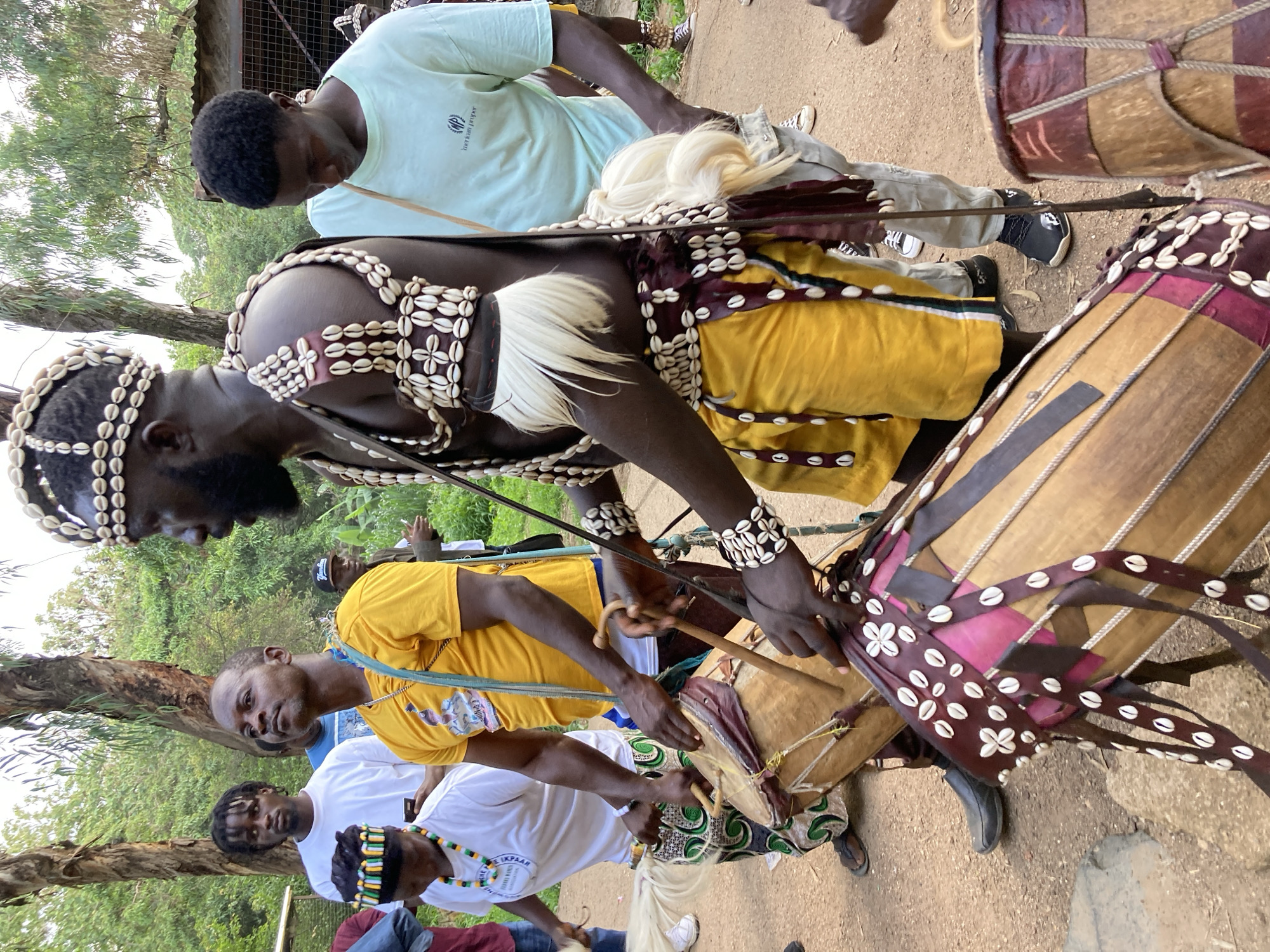An outdoor, medium shot captures a group of people engaged in drumming, possibly during a cultural event. The focus is on three men in the foreground, two of whom are actively playing drums. One man, positioned centrally and closest to the viewer, wears a vibrant yellow t-shirt and has short, dark hair. He is looking upwards and to his left, holding drumsticks. To his left, another person, wearing a white t-shirt and a decorated headband, is also playing a drum. This individual's head is bowed, and they are playing with drumsticks. To the right of the central figure, a third man, dressed in traditional attire including a headdress adorned with cowrie shells, a sleeveless top, and yellow shorts, is also playing a drum. He is looking towards the right. Several drums are visible, some with decorative elements like shells and fabric. The background shows a lush green environment with trees and some structures, suggesting a setting within the city of Jos, Nigeria. The ground appears to be dirt or gravel. The overall mood is energetic and celebratory.