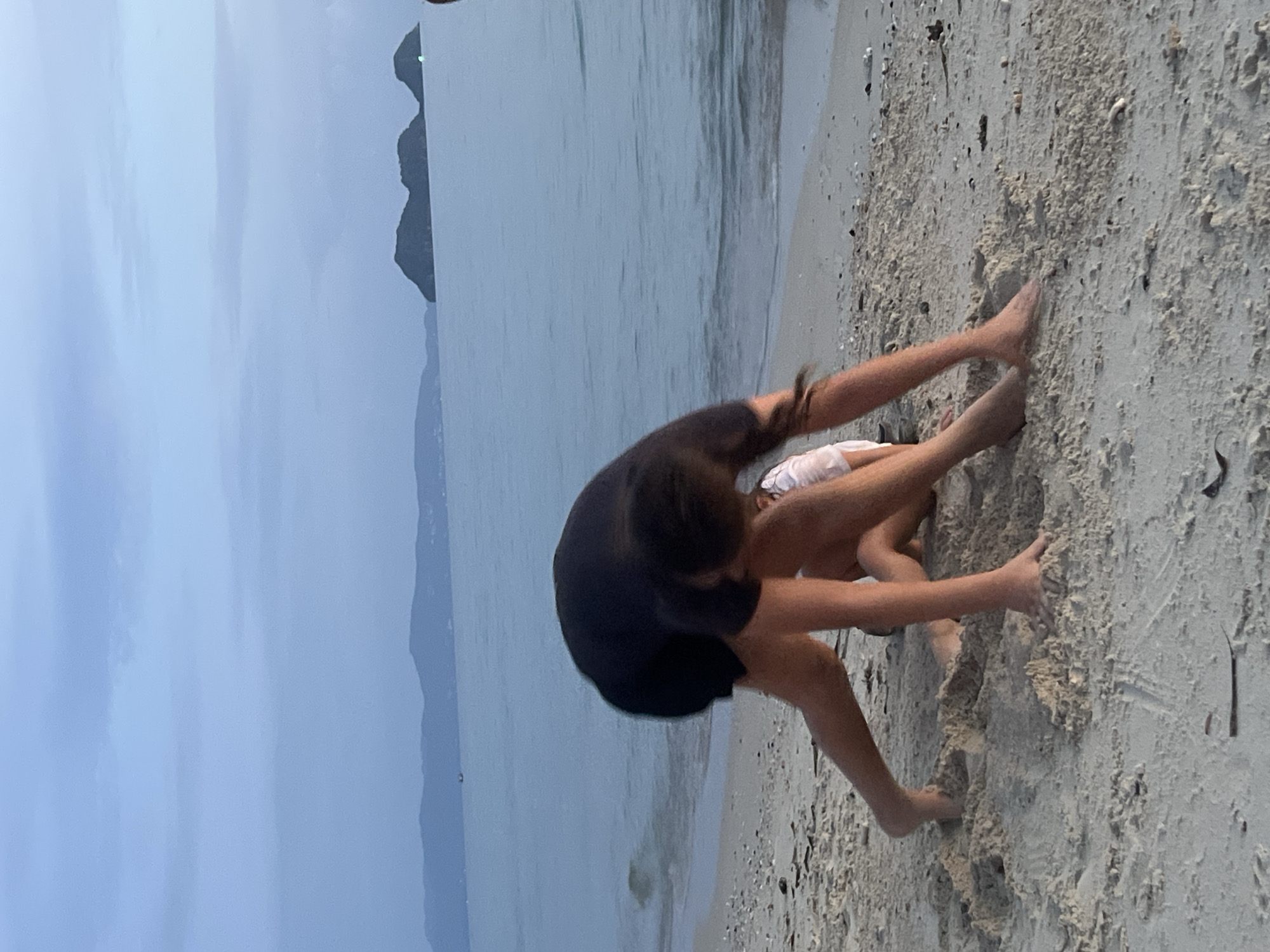 The image shows two children playing on a sandy beach next to the ocean. One child, in the foreground, is wearing a black t-shirt and dark shorts. They are leaning forward, with their arms extended and hands on the sand, appearing to be in motion, possibly jumping or diving. Their legs are bent, and their feet are visible on the sand. The second child is partially visible behind the first, wearing white shorts. The scene is set on a beach with wet sand in the foreground and the ocean stretching out beyond. The water is calm and reflects the overcast sky. In the background, distant islands or landforms can be seen rising from the sea. The sky is a muted, cloudy gray, suggesting it might be early morning, late evening, or a generally overcast day. The overall mood is playful and energetic, with the children engaged in an activity on the beach. The location cues, such as the islands in the distance and the type of beach, are consistent with a tropical setting like Ko Pha-ngan, Thailand. There is no visible text in the image.