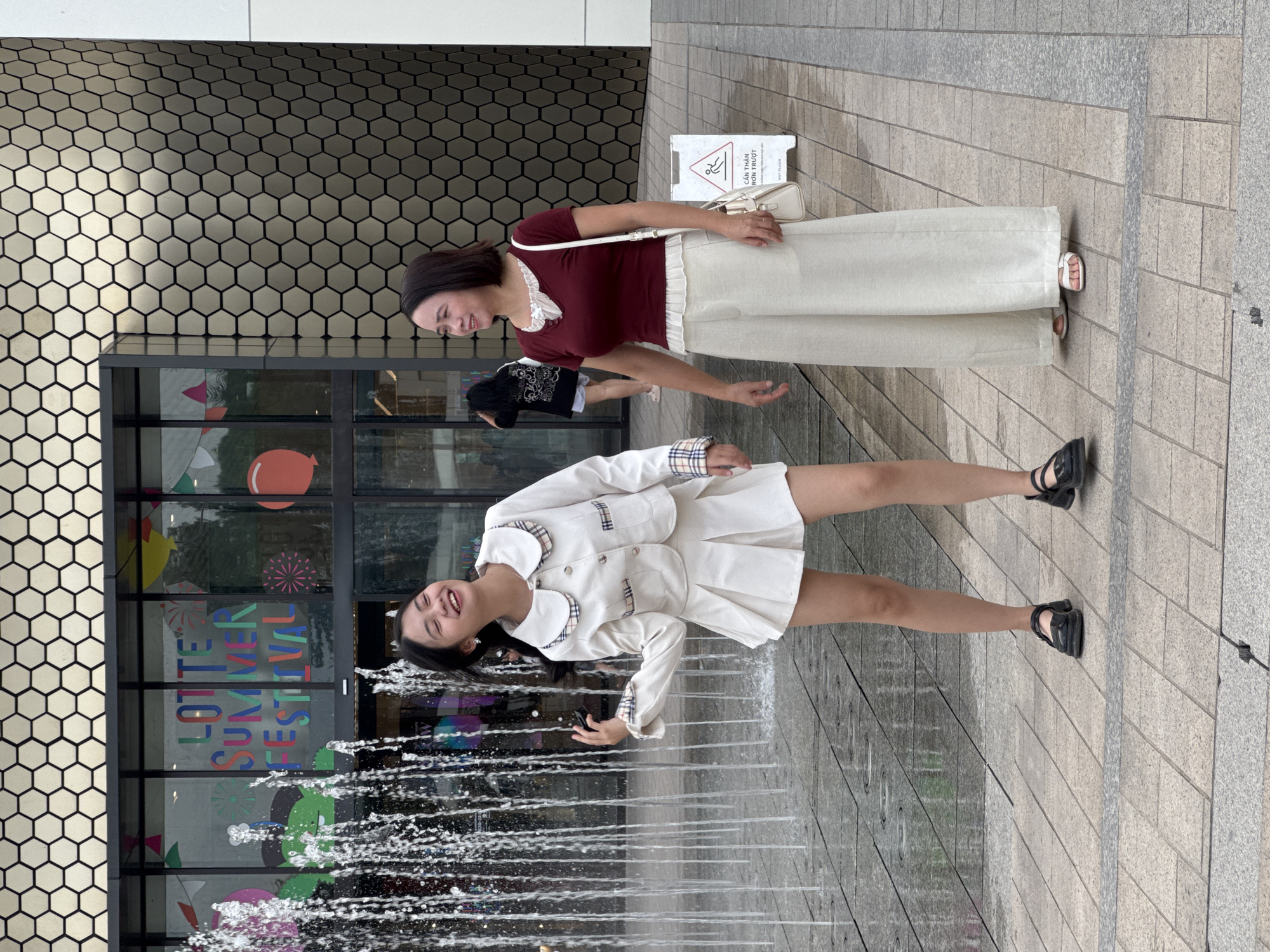 Two women are standing on a paved area in front of a building with a distinctive hexagonal facade. To the left, a water fountain sprays water. The building's windows display colorful text that reads "LOTTE SUMMER FESTIVAL". The woman on the left is young, wearing a white jacket and skirt set with plaid trim and black shoes. She is smiling and looking to her left, with her left arm raised. The woman on the right is older, wearing a maroon top and wide-legged cream pants with white sandals. She has a white crossbody bag and is looking to her left. A wet floor sign is visible to the right of the woman on the right. The scene appears to be outdoors in a public urban space. Based on the "LOTTE SUMMER FESTIVAL" signage and the building's architectural style, the location is likely a Lotte department store or shopping complex in Hanoi, Vietnam.
