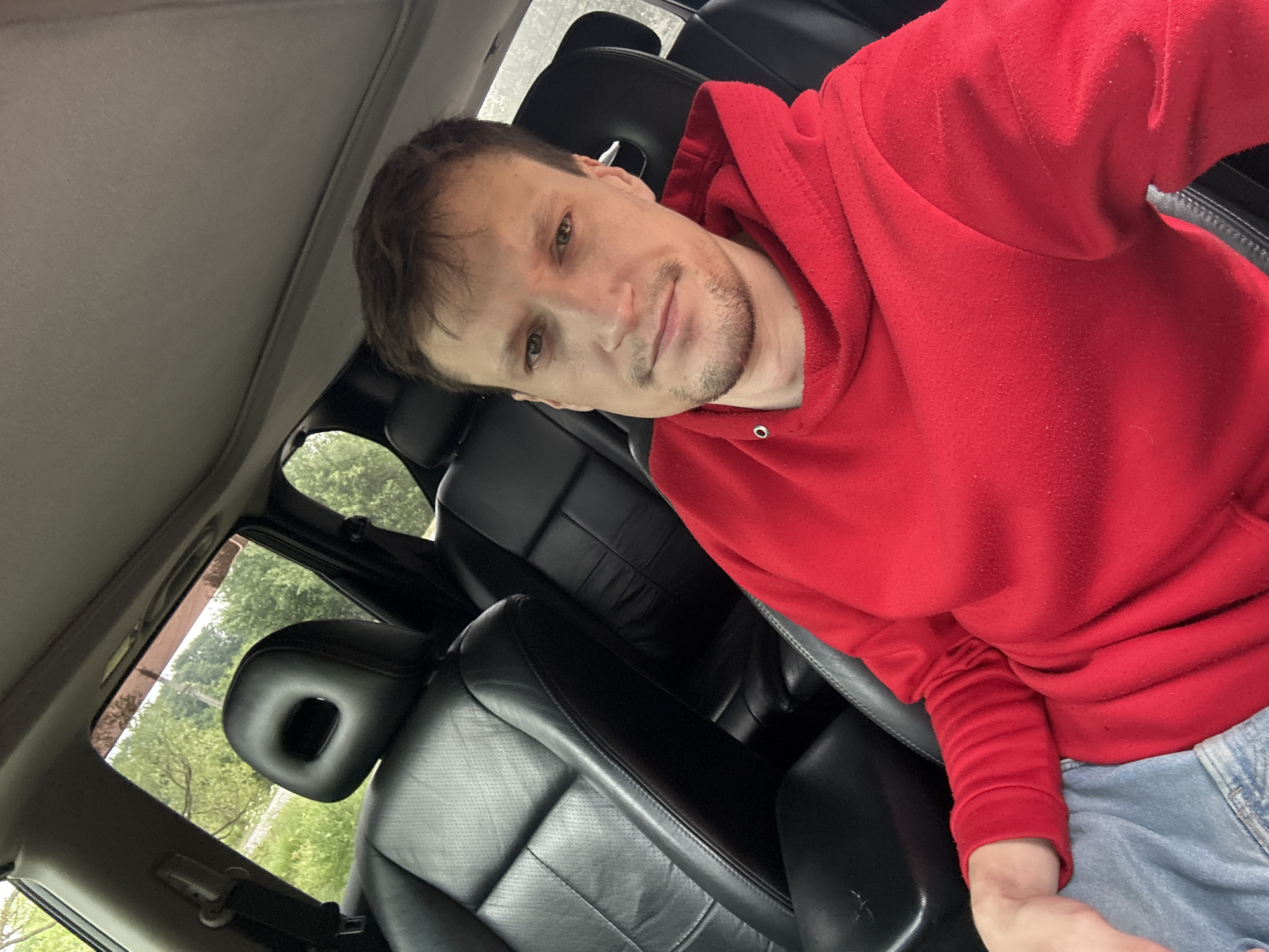 A man is taking a selfie from the driver's seat of a car. He is wearing a red hooded sweatshirt and blue jeans. The car's interior is visible, with black leather seats and a sunroof. Outside the car, trees and greenery can be seen through the windows. The man appears to be in his late 20s or early 30s, with short brown hair and stubble. He has a neutral expression on his face. The location is the city of Makiv, Ukraine.