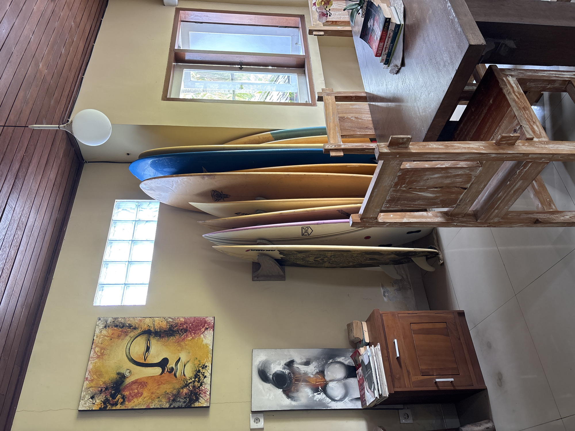 The image depicts an interior scene, likely in Pekutatan, Indonesia, showcasing a collection of surfboards stored against a wall. Several surfboards of varying colors and designs are stacked horizontally. The wall is adorned with two paintings, one featuring a stylized human face and the other a black and white abstract design. A small wooden cabinet is also present, adjacent to the surfboards. Above the surfboards, there is a window and a ceiling light fixture. A wooden table and chair are positioned in the room, partially obscuring the view.