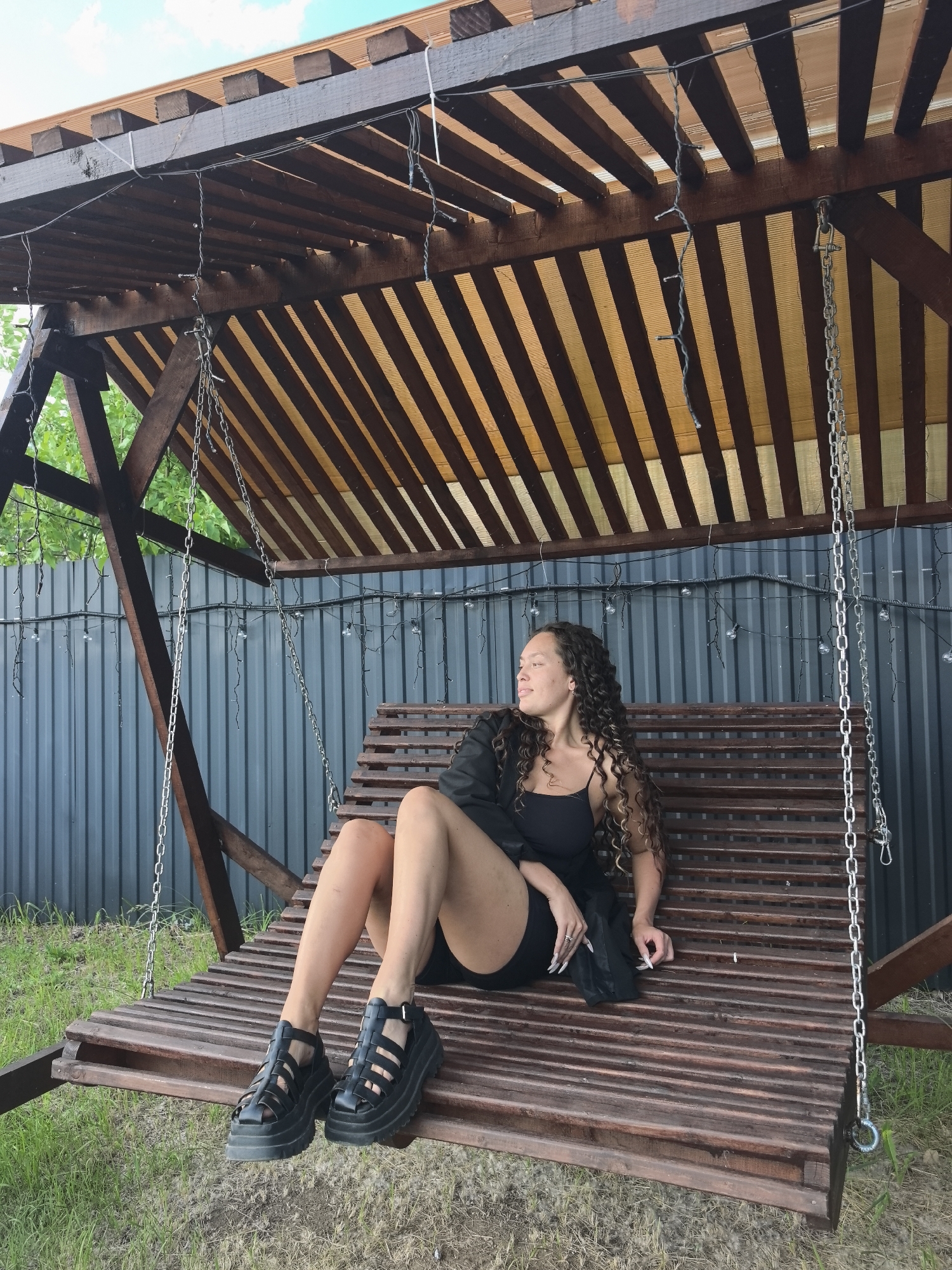 A young woman with long, curly brown hair is sitting on a wooden swing under a wooden pergola. She is wearing a black tank top, black shorts, a black jacket, and black platform sandals. The swing is suspended by chains. Behind the swing is a dark gray corrugated metal fence. There are string lights hanging from the pergola and along the fence. Green grass and some brown patches are visible on the ground. The sky is blue with some clouds. The location appears to be a backyard or an outdoor seating area.