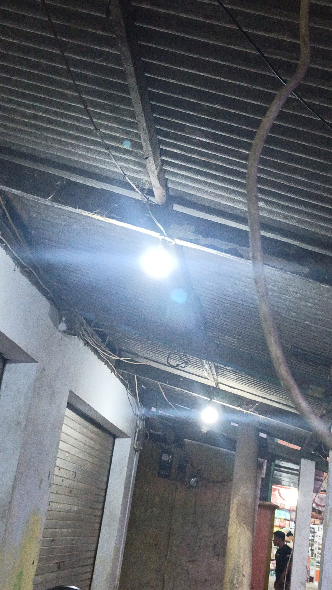 The image shows a dimly lit interior space with a corrugated metal roof supported by wooden beams. Two bright overhead lights are visible, one with a strong glare. Exposed electrical wires are strung across the ceiling. To the left, a closed metal rolling door is visible against a concrete wall. In the background, a person in a dark shirt stands to the right, looking away from the camera. The environment appears to be an outdoor market or a workshop, possibly in a developing urban area, given the rustic construction and exposed infrastructure.