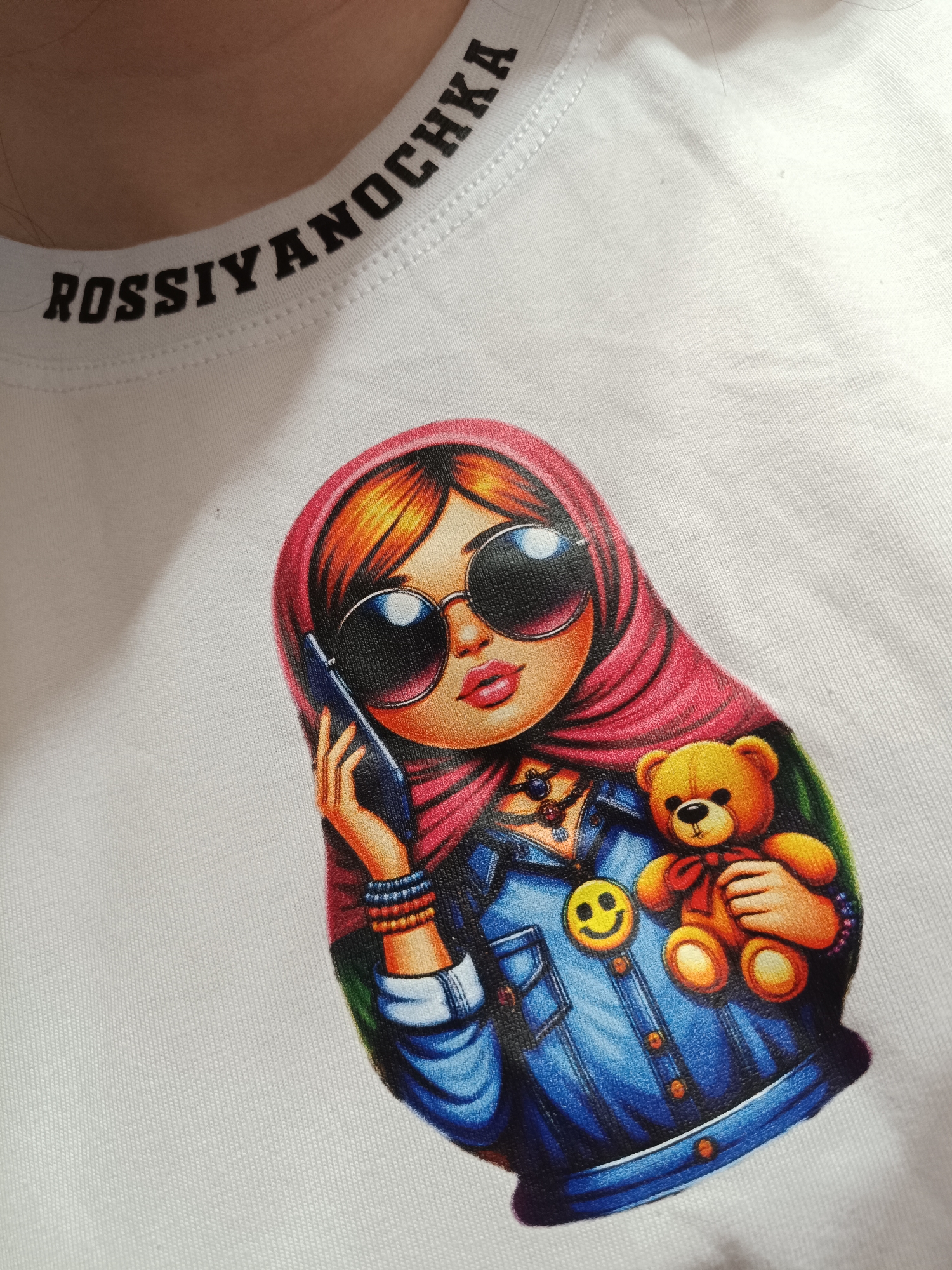 The image presents a close-up view of a white t-shirt worn by a person, whose neck and shoulder are partially visible, set in Tobolsk, Russia. The t-shirt features a prominent, vibrant graphic of a stylized female cartoon character, shaped like a matryoshka doll. The character has bright orange hair peeking from under a red hijab. She wears large, round, reflective sunglasses and a blue denim jacket, accessorized with a multi-strand necklace, beaded bracelets, and a yellow smiley face pin. With her right hand, she holds a black smartphone to her ear, while her left arm cradles a small, light brown teddy bear with a red bow. The t-shirt's ribbed collar displays black, uppercase text that reads "ROSSIYANOCHKA." The graphic's design blends traditional Russian symbolism with modern elements, conveying a youthful and contemporary feel. No specific activity beyond wearing the shirt is depicted by the person, but the character appears to be mid-conversation.