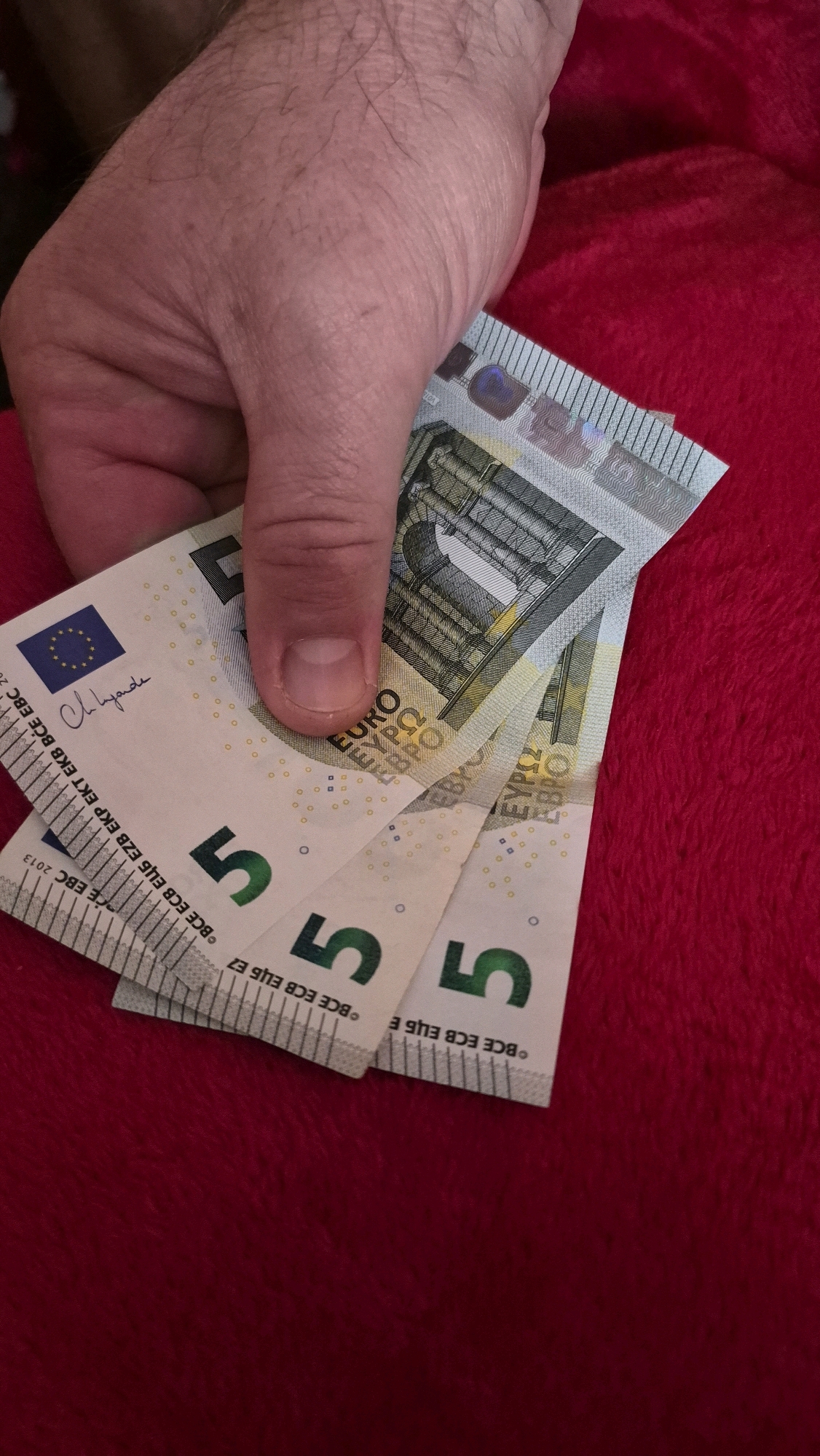 A close-up shot shows a Caucasian hand holding three 5 Euro banknotes against a textured, dark red background. The banknotes are fanned out, with the numbers "5" clearly visible on each. The hand, with visible pores and fine hairs, grips the bills with a thumb and forefinger. A watermark of a signature and a European Union star emblem are visible on the leftmost bill. The banknotes appear to be new or in good condition. The overall lighting suggests an indoor setting. There is no indication of the city of Krauja, Latvia in the image.
