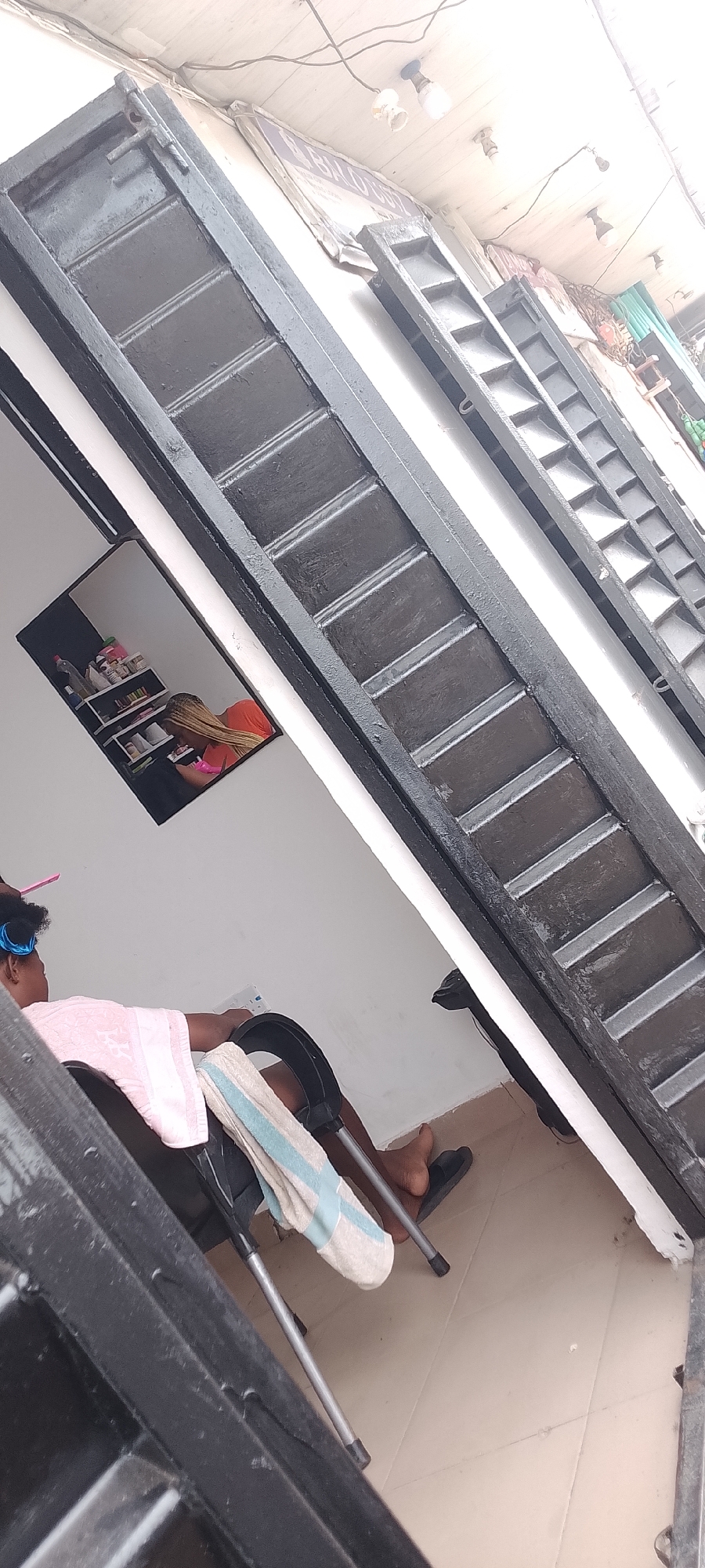 In Ado, Nigeria, a person sits in a black salon chair, their hair styled with blue rollers and a pink comb, draped in a pink and white towel. Next to them, a white and blue striped towel rests on the chair. Their feet are bare, clad in black flip-flops, on a tiled floor. A mirror on the wall reflects a shelf unit with various items. Above, the ceiling is adorned with light fixtures and wires, suggesting a commercial establishment. The scene is framed by dark, slatted metal structures.