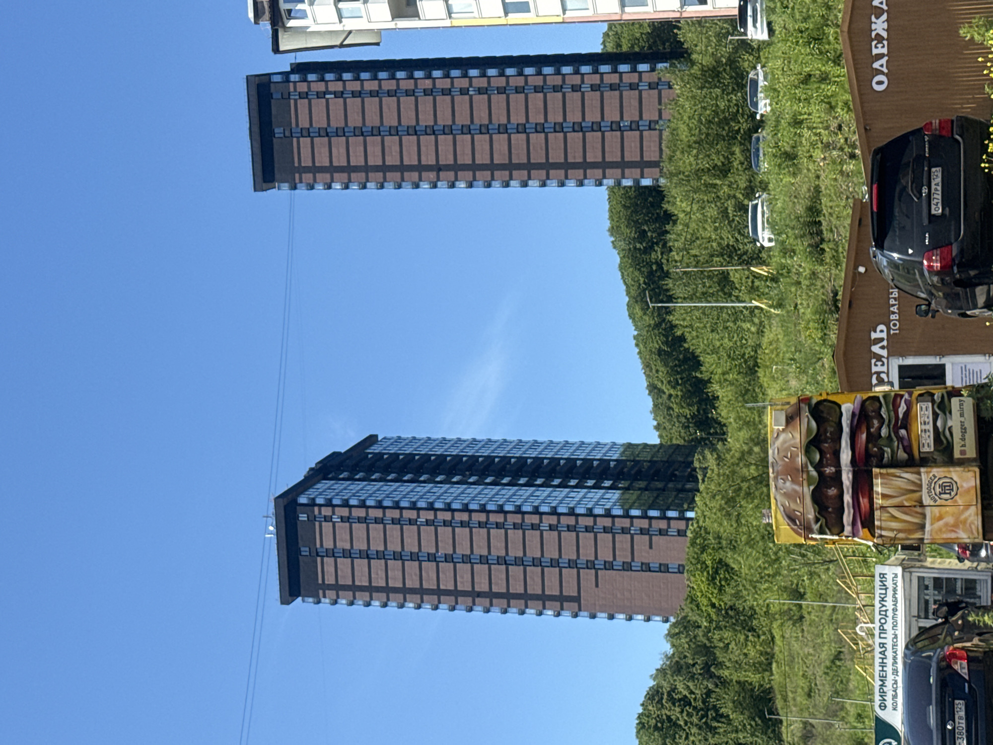 The image captures a street scene in Vladivostok, Russia, dominated by two tall, modern apartment buildings with brown brick facades and dark tinted windows. These buildings are situated behind a lush, green, tree-covered hillside, suggesting a somewhat elevated or natural setting within the urban landscape.
In the foreground, there is a brightly painted food truck featuring a large, detailed mural of a hamburger and french fries. The truck is yellow and appears to be a mobile vendor. To the left of the food truck, a small kiosk with a sign reading "ФИРМЕННАЯ ПРОДУКЦИЯ" (Brand Products) and listing "КОЛБАСЫ-ДЕЛИКАТЕСЫ-ПОЛУФАБРИКАТЫ" (Sausages-Delicacies-Semi-finished products) is visible. Several vehicles are parked near the kiosk and food truck, including a black car with a license plate that appears to read "C380TB 125" and another black SUV with a license plate "0477PA 125".
Behind the food truck and kiosk, there are several more low-rise commercial buildings with brown wooden facades, one of which has signage that includes the word "ТОВАРЫ" (Goods) and "ОДЕЖА" (Clothing). On the hillside behind these structures, several white cars are parked. The sky is clear and blue, indicating a sunny day. The overall impression is a blend of modern residential architecture with the commercial and everyday life of a street-level view.