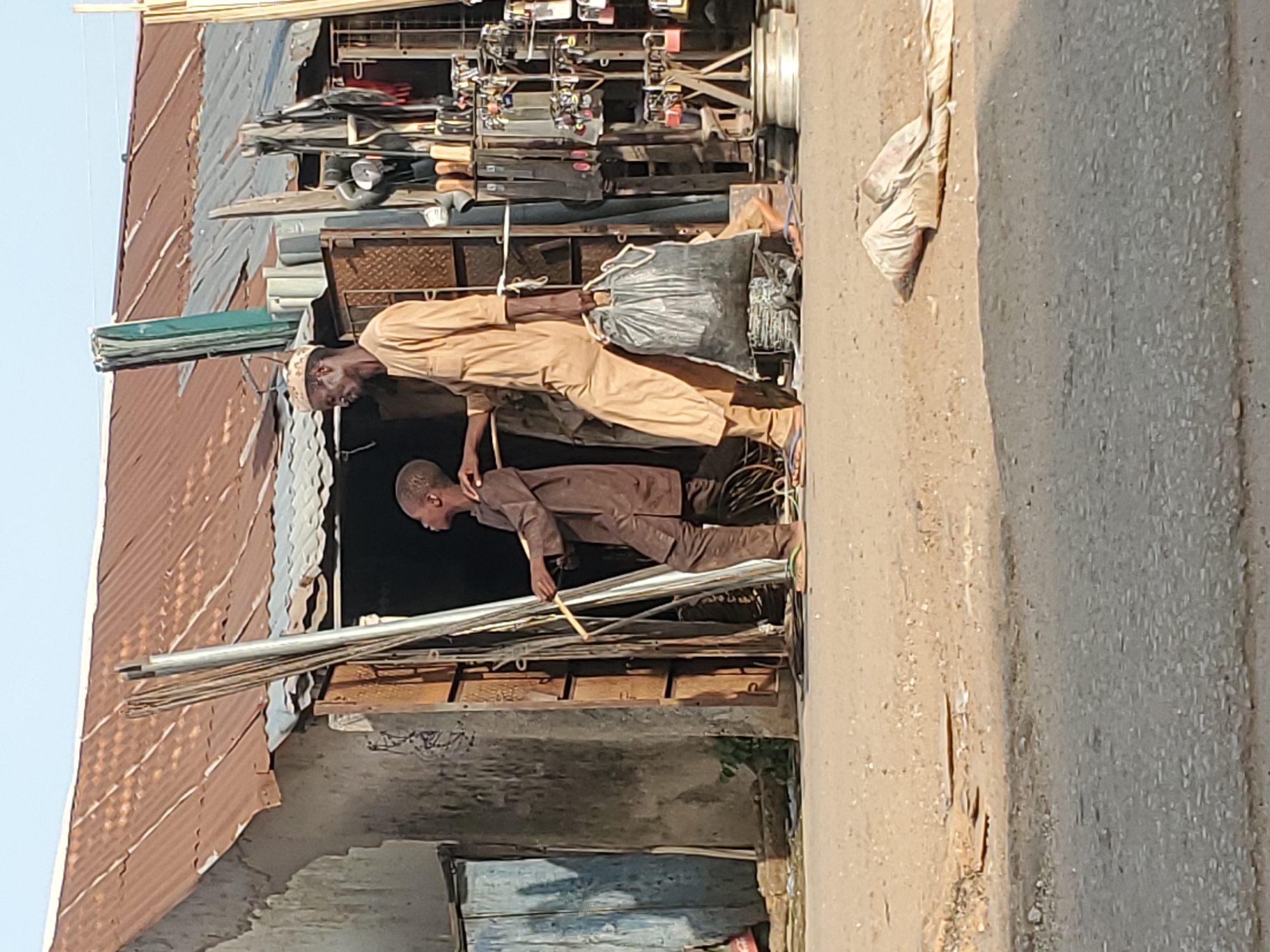 In Mararaba, Nigeria, an eye-level, outdoor shot depicts two males standing in front of a building with a corrugated metal roof. The man in the foreground is wearing a brown, long-sleeved top and trousers. The man behind him wears a tan, long-sleeved top and trousers and a white prayer cap. A black bag hangs from his shoulder. Silver metal rods protrude from the building in front of him. Shelves full of assorted items are visible behind the tan-clad man. The unpaved ground transitions into a paved road on the right.