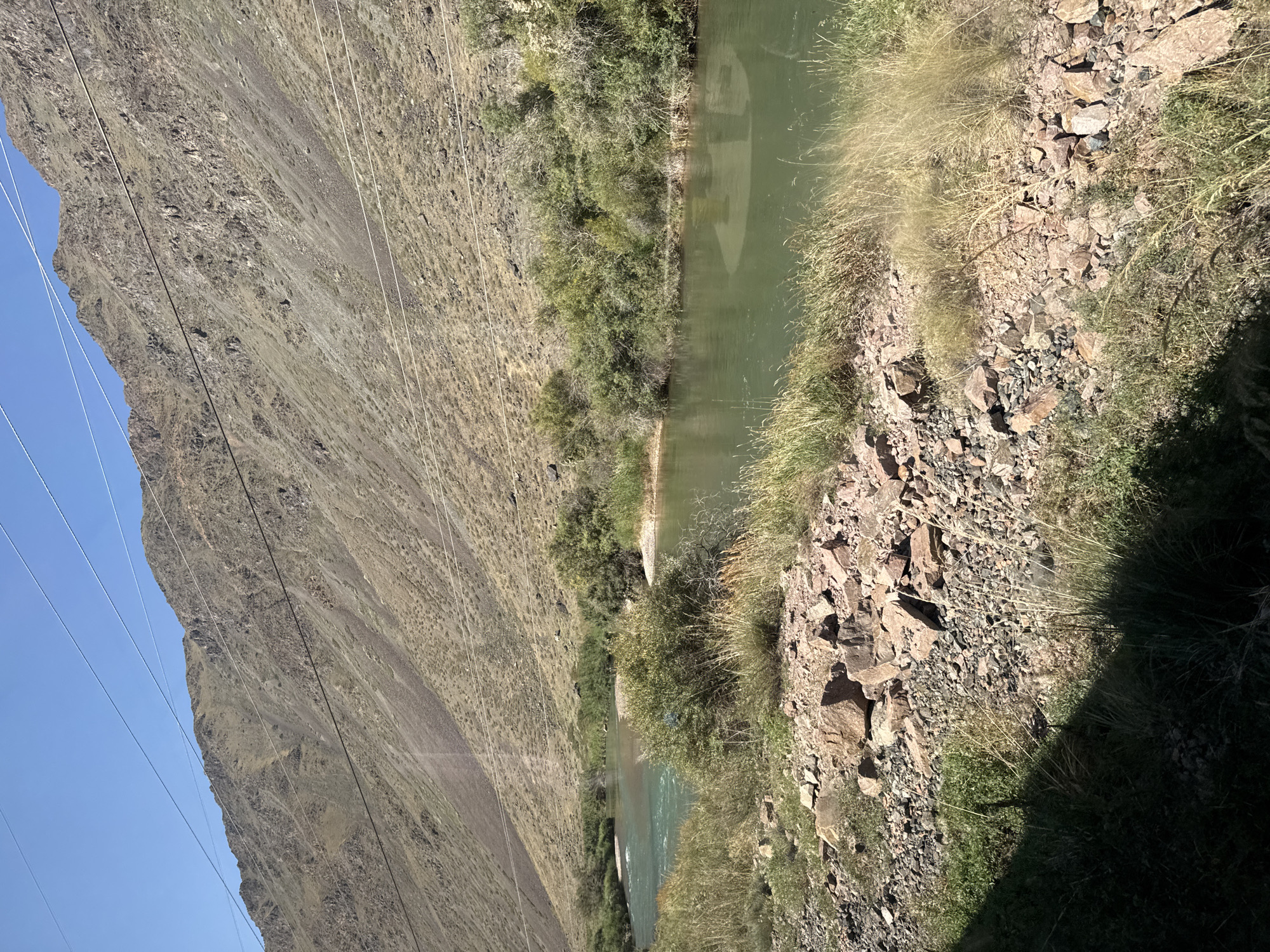 The image depicts a wide, green river flowing through a valley. On the right bank of the river, dense green trees and bushes grow along the water's edge. The foreground on the right consists of dry, tall grass and a rocky, pebbly shoreline with some larger, reddish-brown rocks scattered amongst the smaller stones. In the background, a steep, barren mountain slopes upwards towards a clear blue sky. Several power lines are strung across the upper portion of the mountain and sky. The environment appears dry and somewhat arid, with the exception of the lush riverbank vegetation. There are no people or discernible structures in the image. The scene is devoid of any activity, presenting a natural, untouched landscape. The overall impression is one of remote natural beauty. It is possible this image was taken near Krasnyy Most, Kyrgyzstan, a location known for its scenic river valleys and mountainous terrain.
