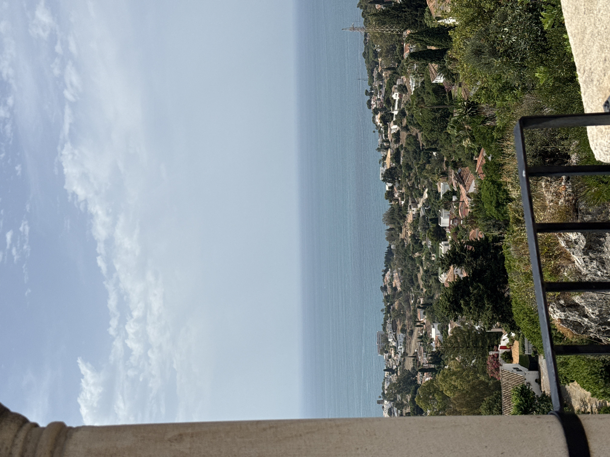The image shows a panoramic view overlooking the coastal city of Benalmádena, Spain. In the foreground, a stone column and a black metal railing frame the scene. The middle ground is filled with a dense, green landscape of trees and vegetation, interspersed with numerous white-roofed houses and buildings with terracotta tiles, indicative of Mediterranean architecture. A winding road is visible snaking through the hillside. Beyond the cityscape, the vast expanse of the bright blue Mediterranean Sea stretches to the horizon under a hazy, pale blue sky with scattered white clouds. The lighting suggests it is daytime, likely late morning or early afternoon. No people or animals are visible in the image.