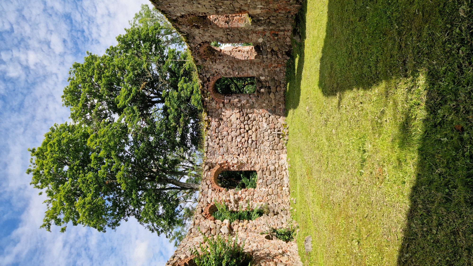 The image shows the ruins of a stone building with arched windows. The walls are made of rough-hewn stones and bricks. Green grass covers the ground inside and outside the ruins. Trees with lush green foliage are visible behind the walls, and a partly cloudy blue sky is in the background. The scene appears to be a historical site. This is a view of the Embūte Castle Mound ruins in Embūte, Latvia.