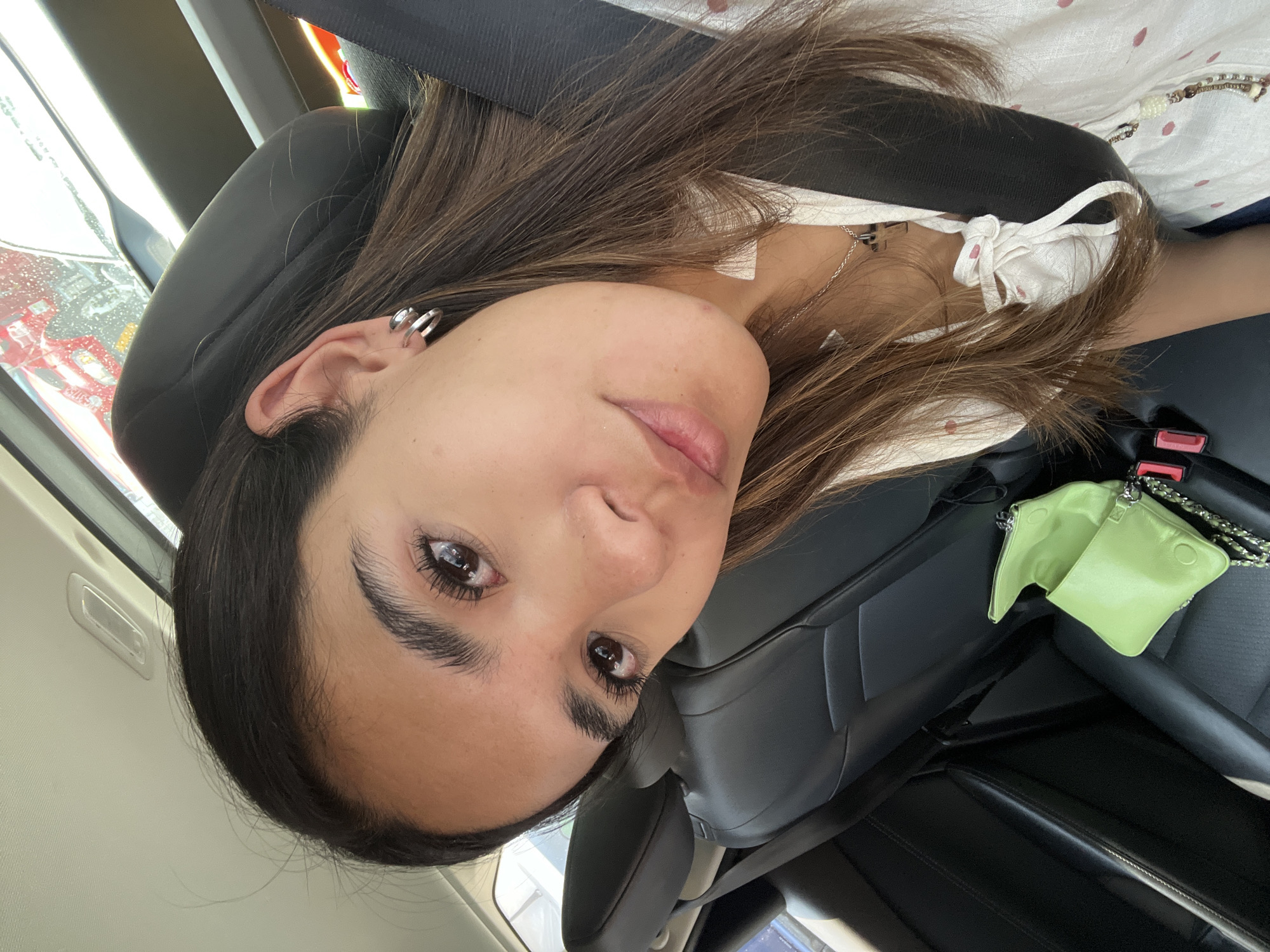 A woman sits in a car, buckled in with a seatbelt. She appears to be the only occupant visible. She has long, brown hair and is wearing silver hoop earrings. She has on a white top with pink polka dots and a knotted tie at the neckline. A small, light green purse rests on the black leather passenger seat. Through the car window, a rainy cityscape is visible, potentially within Mexico City, Mexico.