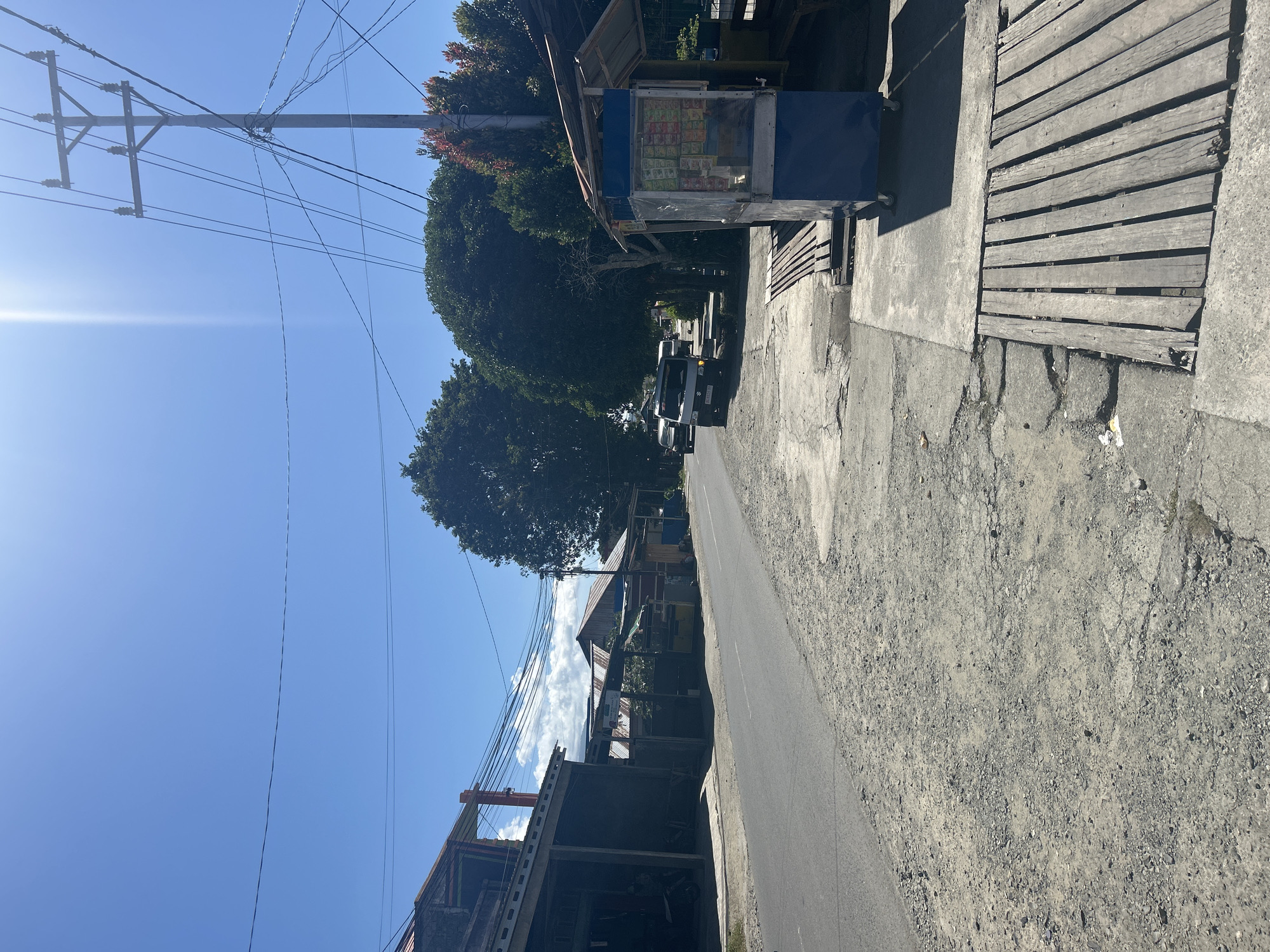 A bright, sunny day unfolds in Mandaong, Indonesia, as a two-lane asphalt road cuts through a semi-urban landscape. On the left side of the road, a row of single-story buildings with corrugated metal roofs and open storefronts line the street. A motorcycle is visible inside one of the dark interiors. The road's shoulder is a mix of gravel and cracked concrete.
Across the street on the right, a small blue and white kiosk with a wooden roof stands at the edge of a concrete sidewalk. The kiosk displays various packaged goods. Adjacent to the kiosk, a wooden bench is set up. Further down the sidewalk, another wooden structure, possibly a covered waiting area or stall, is visible. Wooden planks form a pathway over sections of the uneven concrete.
A few vehicles are present: a light-colored van and another car are stopped or moving slowly on the road in the distance. A utility pole with numerous wires stands prominently on the right side of the road, contributing to the overhead network of electrical and communication lines that crisscross the sky. Lush green trees, some with reddish foliage, dot the background, interspersed with the buildings. The sky is a clear, vibrant blue with a few wispy clouds and a strong sun creating a bright lens flare effect.