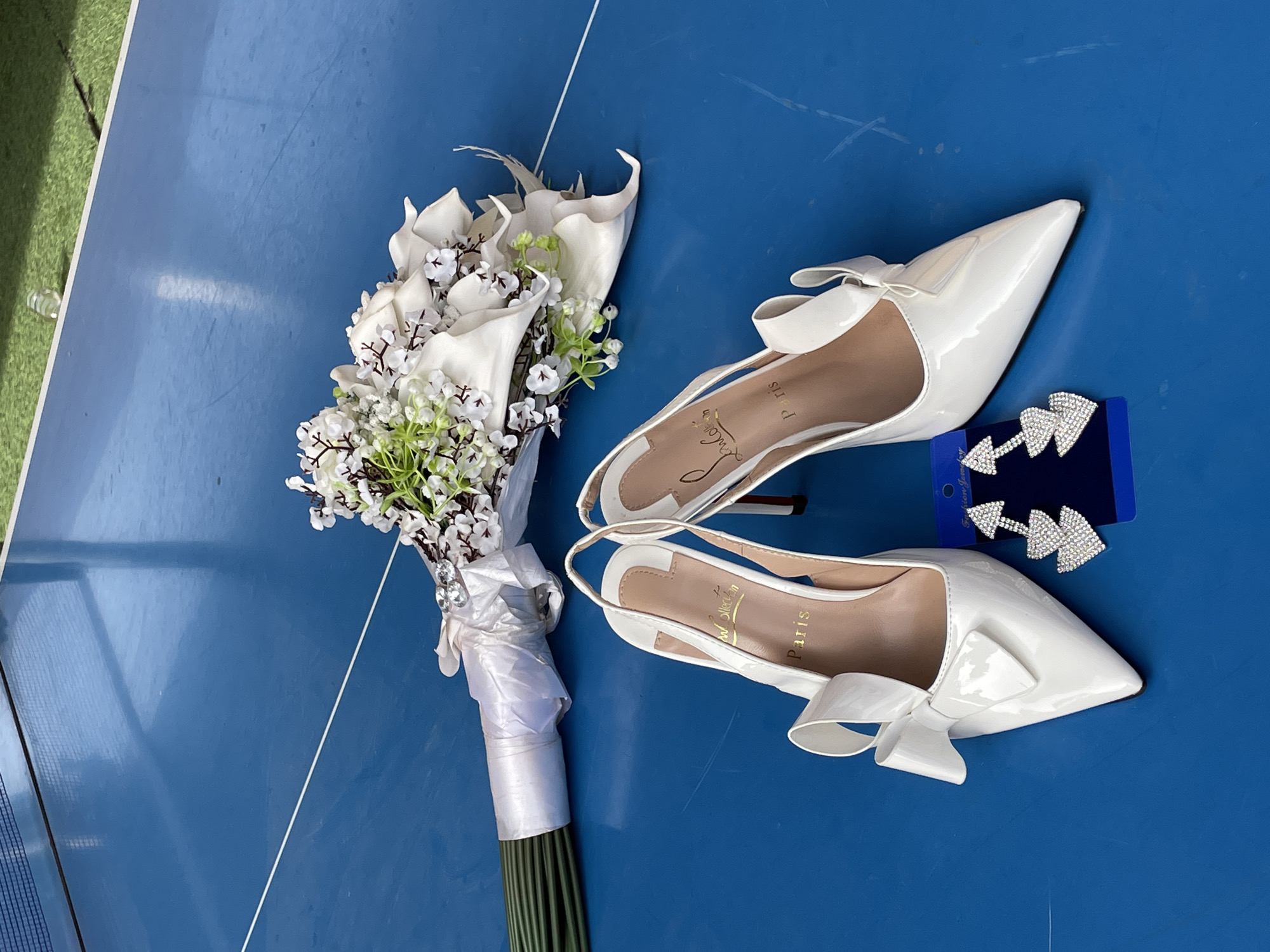 A high-angle shot shows a wedding arrangement on a blue surface. The arrangement includes a bouquet of white flowers and foliage tied with a white ribbon, a pair of white patent leather heels with bows, and a blue card with sparkly arrow-shaped earrings.