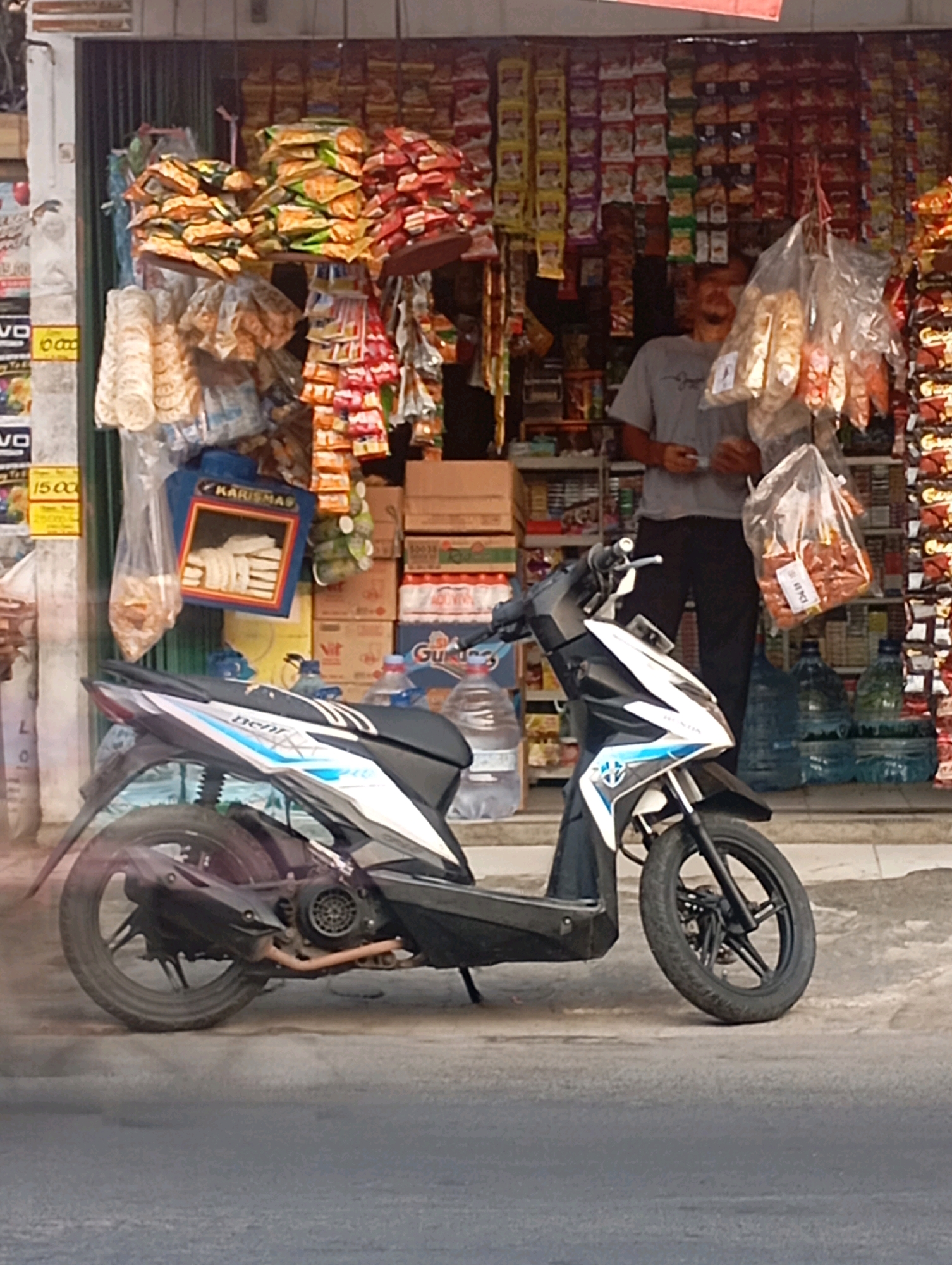 The image depicts a street-level view of a small shop in Babelan, Indonesia, with a motorcycle parked in front. The shop appears to be a convenience store or warung, displaying a variety of packaged snacks and drinks on shelves and hanging from the storefront. Several large plastic bottles of water are visible. A man, partially obscured by hanging merchandise, stands inside the shop, wearing a grey t-shirt and dark pants. A black and white scooter is parked on the pavement in front of the store, angled towards the right. The ground is paved, and a street runs in the foreground, showing a slight haze, possibly from exhaust. The overall scene suggests a typical commercial setting in a local Indonesian neighborhood.