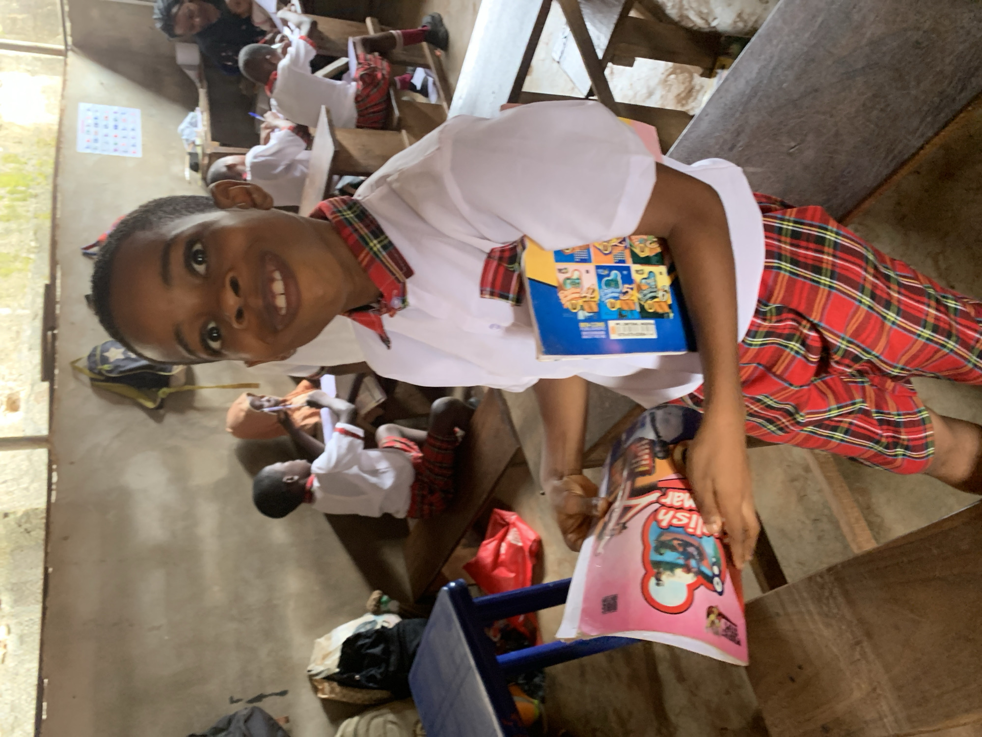 The image shows a classroom in Ukwu-Abwa, Nigeria. A young student in a white shirt with a red plaid collar and red plaid pants is smiling at the camera. He is holding a blue book in one hand and a pink book titled "English Grammar" in the other. Other students, also in white shirts and plaid pants, are seated at wooden desks in the background, appearing to be writing or studying. A calendar hangs on the wall. The room appears to be sparsely furnished and modestly maintained.
