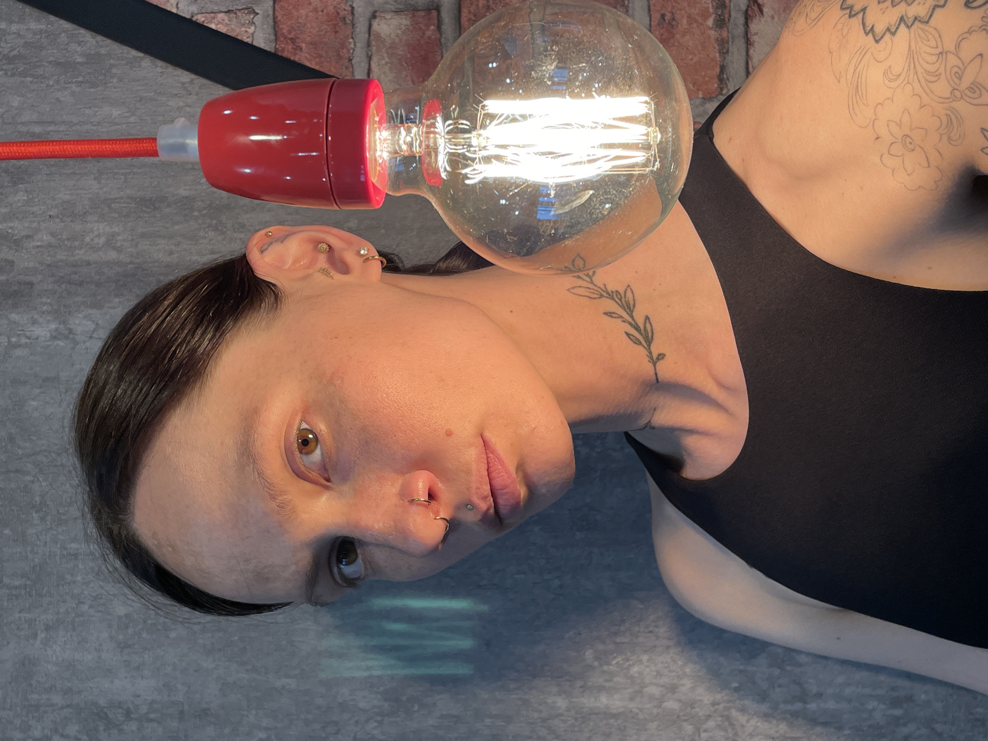 A fair-skinned individual with dark hair, wearing a black tank top, is positioned at an oblique angle relative to the camera, featuring multiple piercings on the left ear, nose, and lip, and tattoos on the neck and right shoulder. The person's gaze is directed slightly downwards and to the right. An illuminated vintage-style light bulb with a red fixture is situated adjacent to the person's head, obscuring part of the neck. The background consists of a grey surface, possibly a floor or wall, and a brick wall is partially visible. The image was not taken in the city of Izhevsk, Russia.