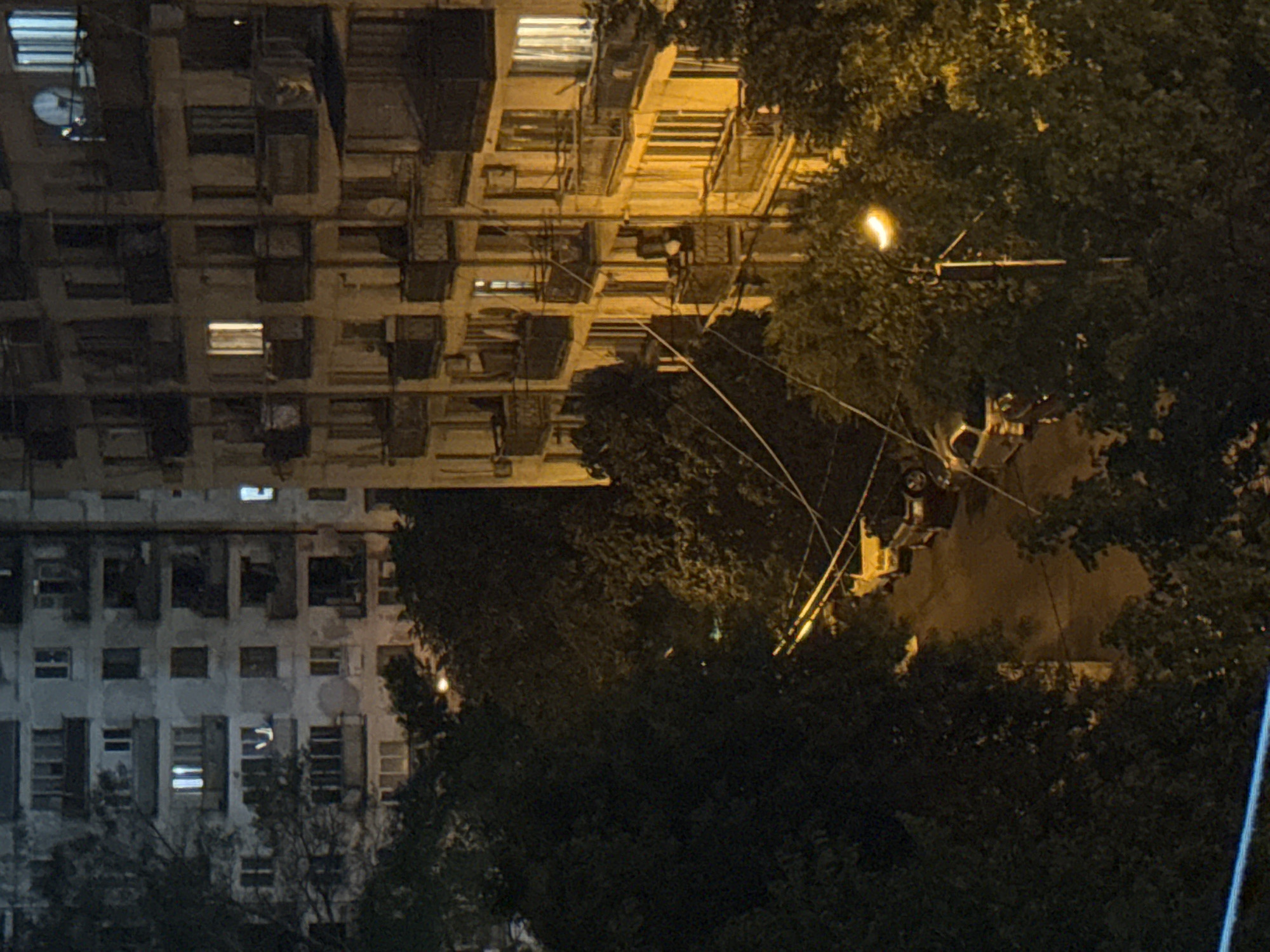 The image depicts a street scene at night in Tbilisi, Georgia, likely in a residential area. Two multi-story apartment buildings, characteristic of Soviet-era architecture with their repetitive balconies and window patterns, dominate the background. One building on the left appears slightly older and darker, while the building on the right is illuminated by warm yellow light from its windows and balconies. Trees with dense foliage line the street, partially obscuring the lower levels of the buildings and casting shadows. The street itself is paved and appears to be a typical urban road. Several cars are parked along the street, including a yellow car and a dark-colored SUV. A street lamp is visible, its warm light illuminating the surrounding trees and a section of the street. Overhead, several power lines or communication cables crisscross the scene, adding to the urban feel. The overall atmosphere is quiet and nocturnal, suggesting a residential neighborhood settling down for the night. There are no people visible in the image.