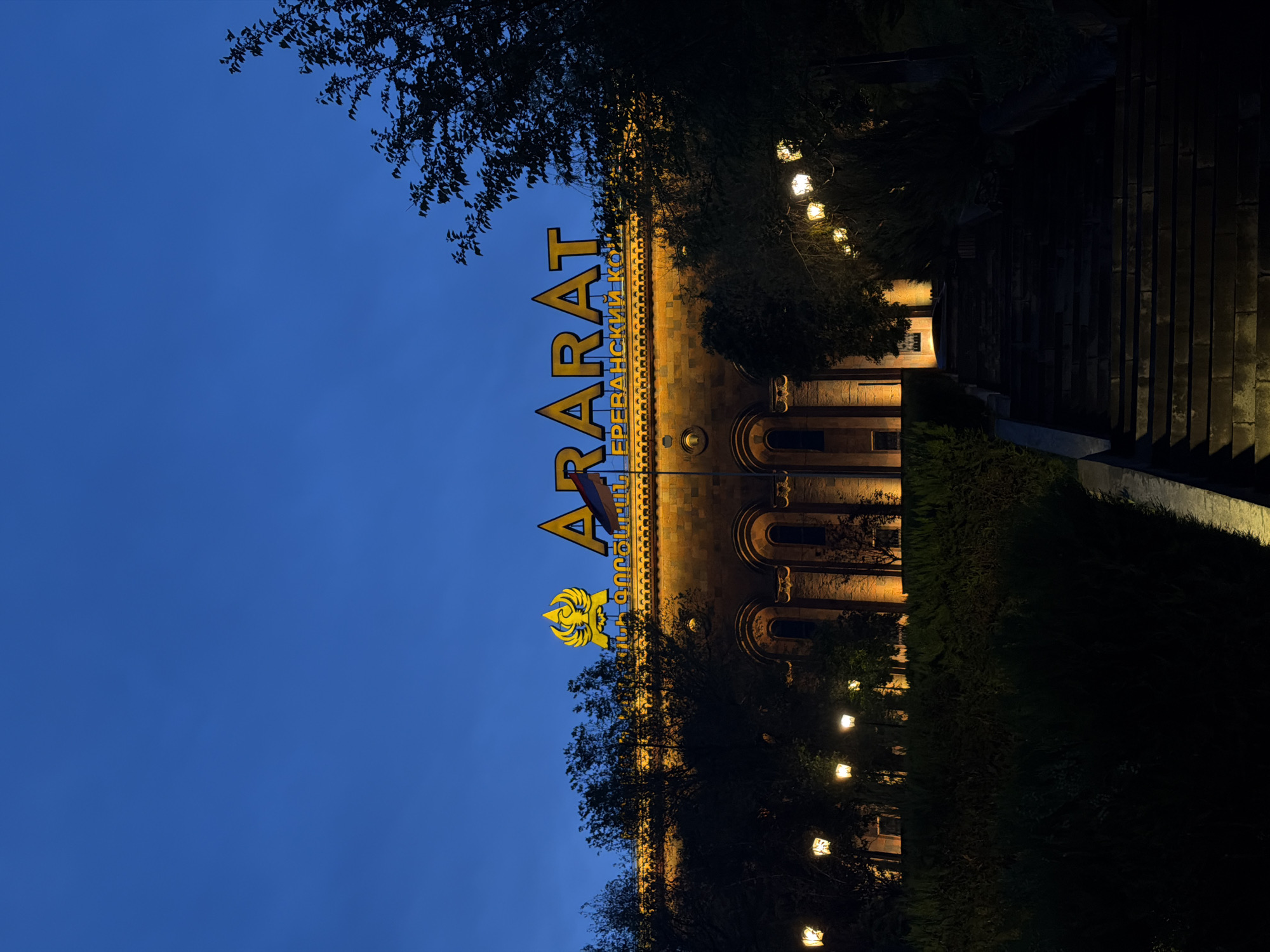 The image depicts the Ararat Brandy Factory in Yerevan, Armenia, at dusk or night. The building has a tan facade and arched windows, illuminated by lights. The "ARARAT" sign in yellow letters is prominent on the building, with additional text in Armenian below it. A flag is visible on the building's facade. Trees and shrubbery surround the building, with strings of lights hanging from the trees. A set of stairs is visible on the right side of the frame. The sky is a deep blue color.