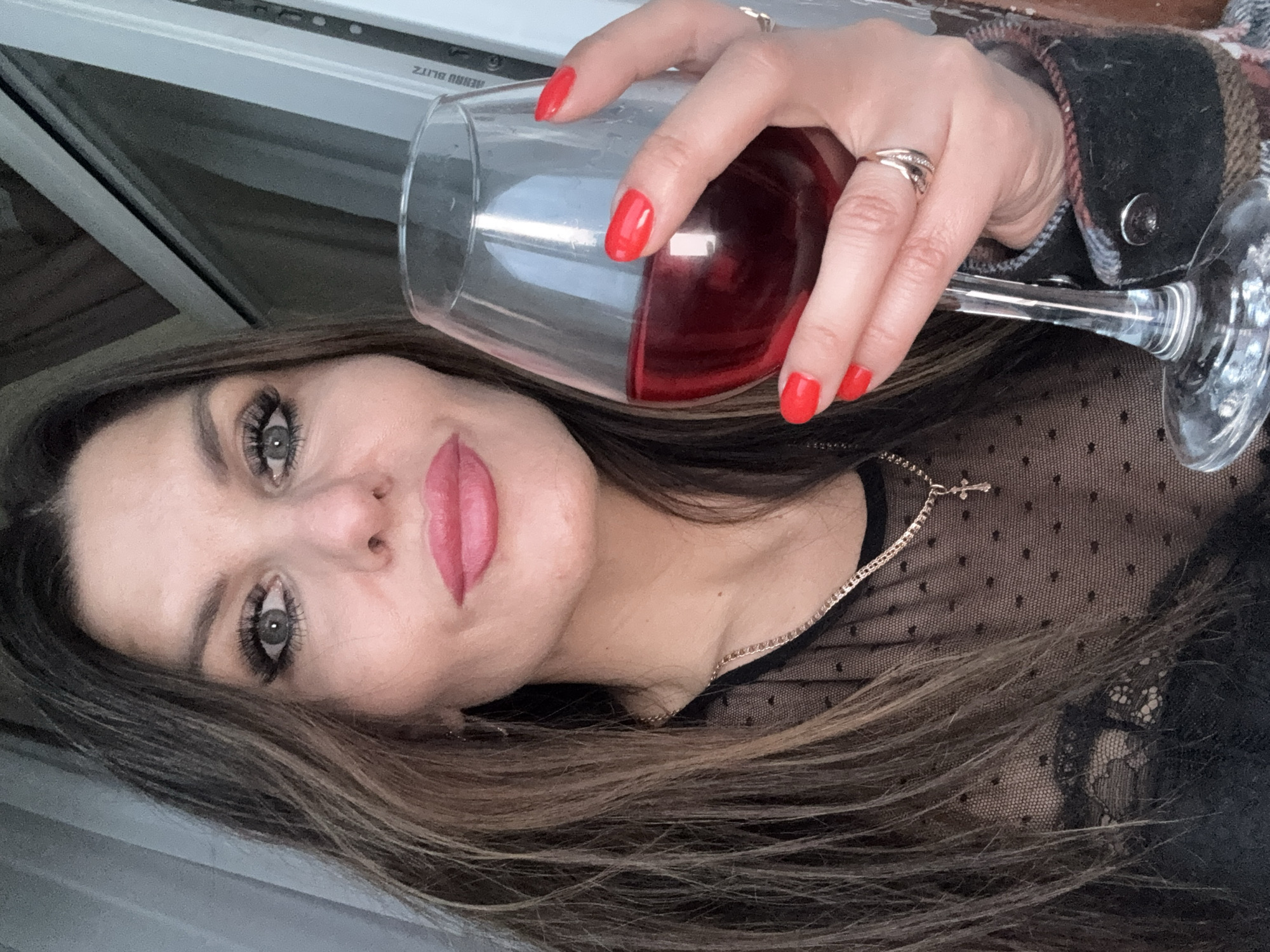 A woman is holding a wine glass filled with red wine. She has red nail polish and several rings on her fingers. She is wearing a black polka-dot top and a cross necklace. The image appears to be taken indoors. I cannot determine the location as Chaikovsky, Russia from the image.