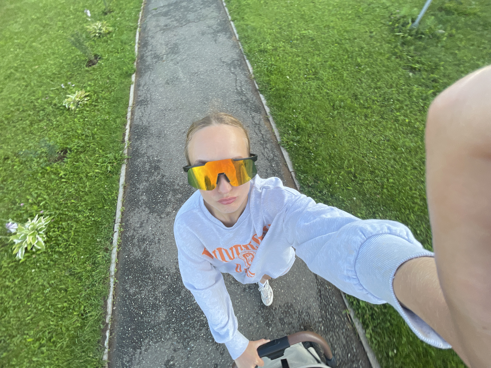 A first-person perspective shot shows a person wearing a white sweatshirt and reflective sunglasses, holding a stroller. The person is standing on a paved walkway next to a grassy area. The scene appears to be outdoors in a park or residential area. The provided location is "Подшивалово, Russia", but this cannot be verified from the image itself.