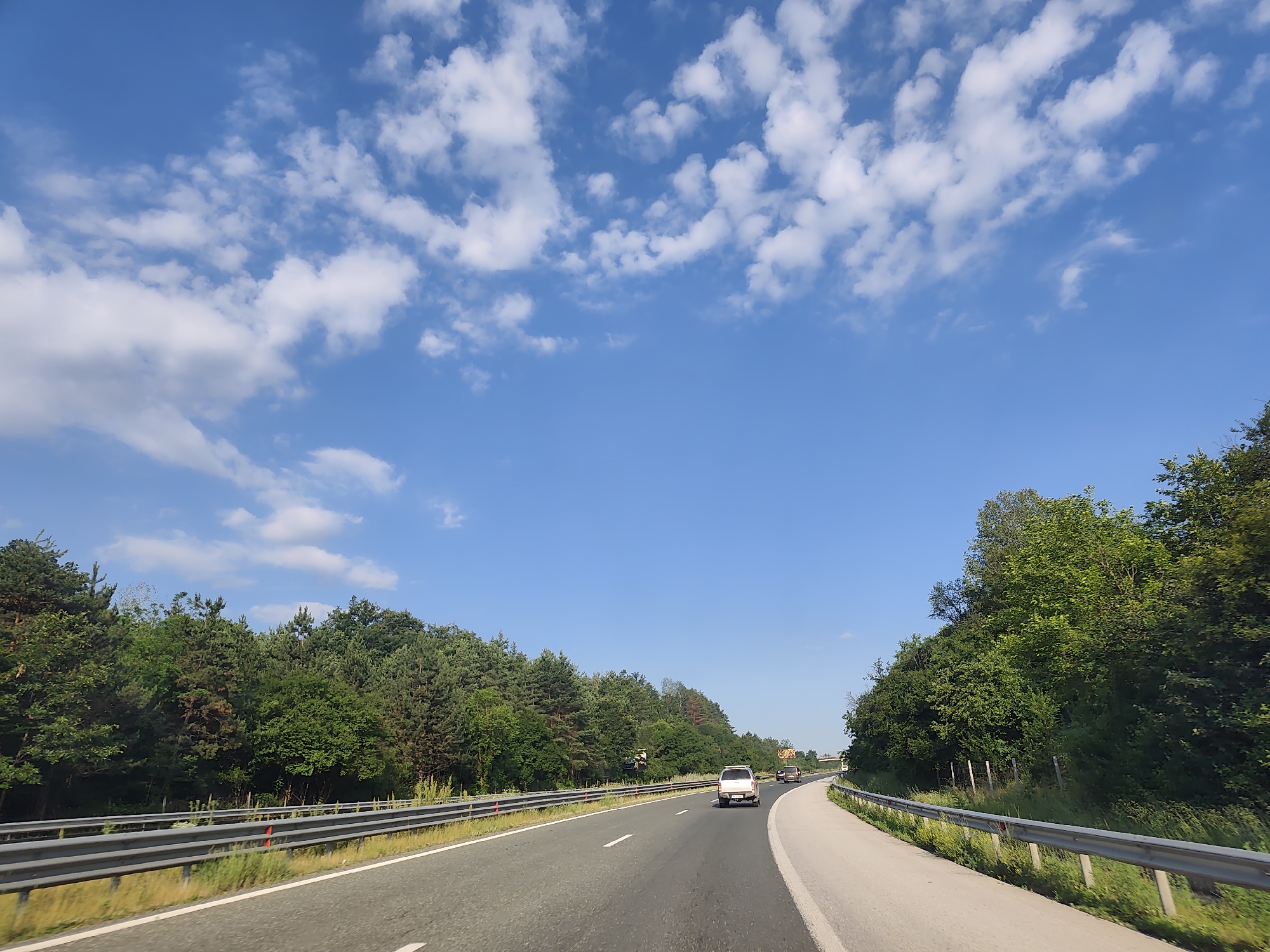 The image depicts a highway scene with a bright blue sky filled with scattered white clouds. The road curves to the right, bordered by lush green trees on both sides. A metal guardrail runs along the edge of the road. A white SUV is driving in the right lane ahead, with another vehicle further down the road. The location is identified as the city of Osikovitsa, Bulgaria.