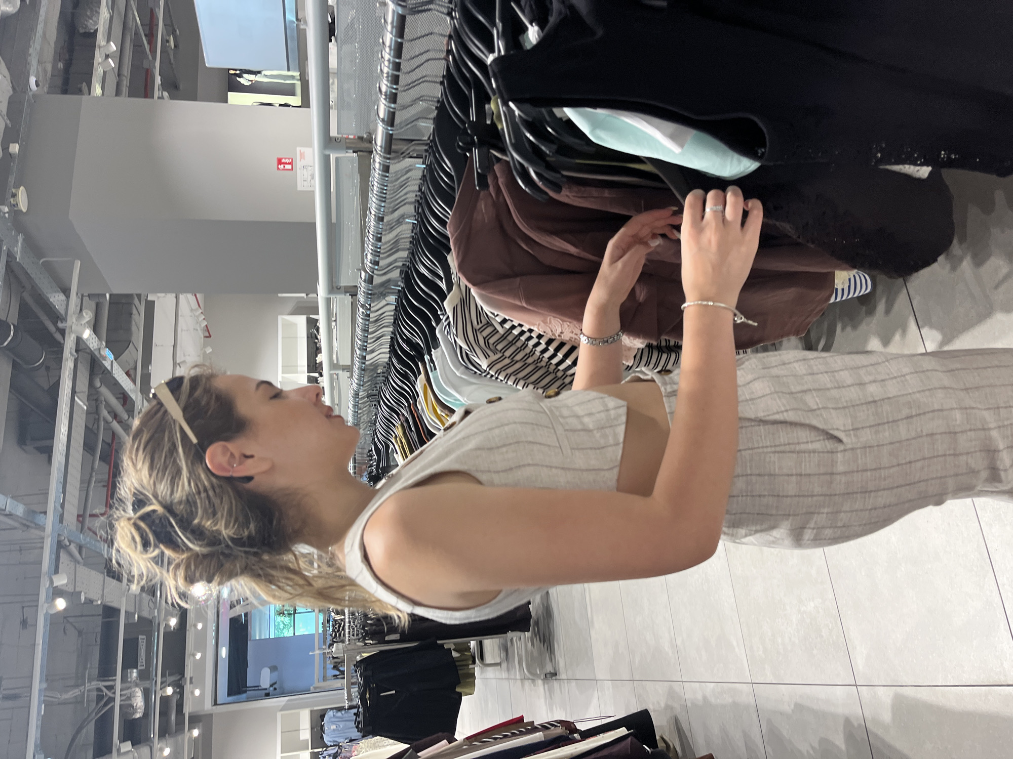 The image captures a young woman browsing clothes in a retail store, potentially located in Blagoevgrad, Bulgaria. She is depicted in profile, facing right, with a slight smile as she actively sifts through a densely packed clothing rack. Her blonde, wavy hair is pulled back, with a pair of sunglasses resting on her head. She wears a light-colored, possibly linen, sleeveless garment with vertical stripes and button details on the bodice. A silver bracelet is visible on her left wrist, and a silver ring adorns her right hand as she reaches for items on the rack.
The clothing rack holds a variety of garments on black hangers, including black tops, a brown item, and striped and white apparel. The setting is an indoor commercial space, characterized by a modern, utilitarian design. The ceiling features exposed pipes, ventilation ducts, and numerous track lights, providing bright artificial illumination. The walls are light gray, and the floor is covered with large, light-colored tiles. In the background, a red fire extinguisher sign is visible on a wall, showing the extinguisher symbol and faint text below it, possibly "EN" and "РУ" (for English and Russian). The overall scene depicts a casual shopping experience within a well-lit, contemporary store environment.