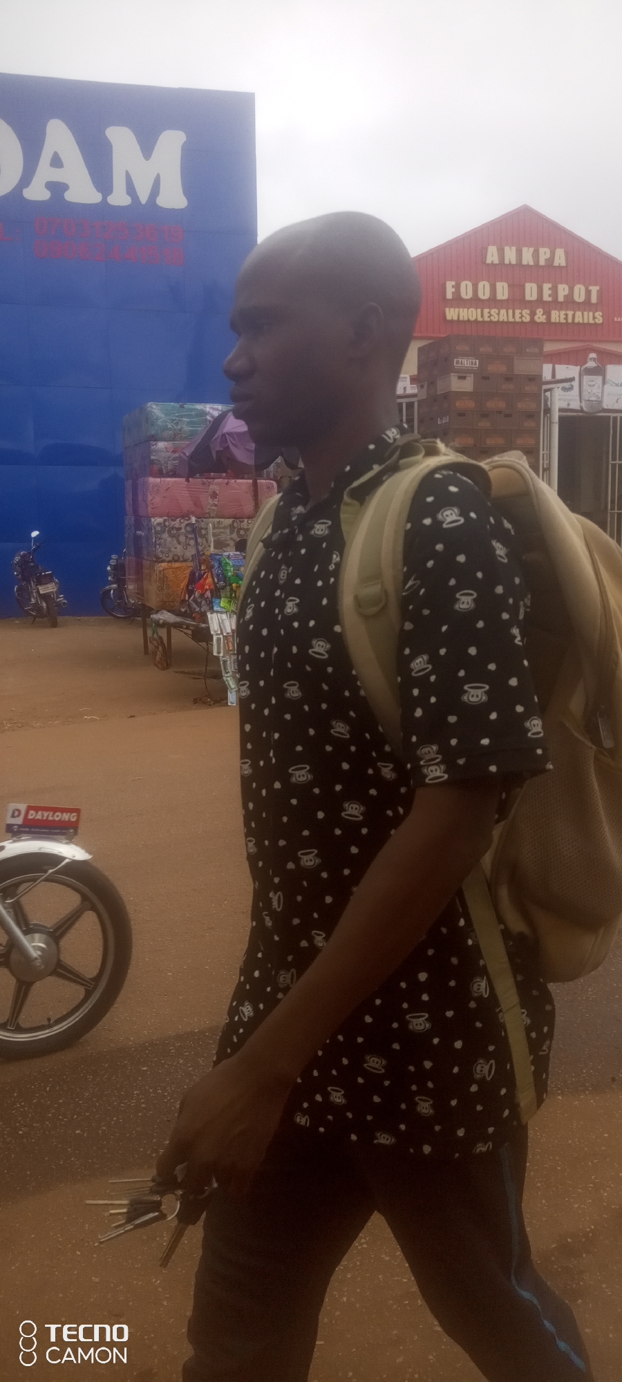 A man in profile walks right, his face turned away from the camera. He wears a black shirt with a white polka dot pattern and a tan backpack. In his right hand, he holds a bunch of keys. He is in front of a building with a red roof that reads "ANKPA FOOD DEPOT WHOLESALES & RETAILS". To the left of the food depot is a large blue sign with white lettering, part of which reads "DAM". Behind the man, there is a stack of mattresses and a small stall with various items. In the background, a motorcycle is parked to the left of the mattresses. The ground appears to be dirt or gravel. The image is taken during the daytime, with an overcast sky. The scene is likely on a street or marketplace in Ankpa, Nigeria, given the visible signage.