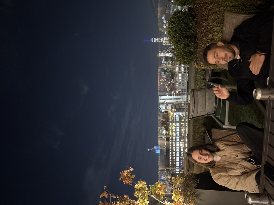 This image captures a nighttime scene on a rooftop terrace in New York City, overlooking the illuminated skyline. The sky is dark with scattered clouds.
Two individuals are seated at a dark-colored outdoor table. On the left, a woman with dark, shoulder-length hair, wearing a light brown quilted jacket over a white top and dark pants, is smiling and looking towards the right. On the right, a man with short dark hair and a beard, wearing a dark long-sleeved shirt, is gesturing with his right hand, his left hand resting on his chest. Three silver beverage cans, identified as Sapporo brand, are visible on the table (two near the woman, one near the man).
The terrace floor is covered with artificial green turf. Potted plants, including one with reddish-orange leaves on the left and green shrubbery in a planter on the right, delineate the seating area. A clear glass barrier separates the terrace from the distant city lights.
The background features a prominent view of the New York City skyline. One World Trade Center, illuminated in blue and white, is visible on the right side of the skyline. Another building with a blue-lit dome is visible towards the center-left. Numerous other city buildings display illuminated windows.