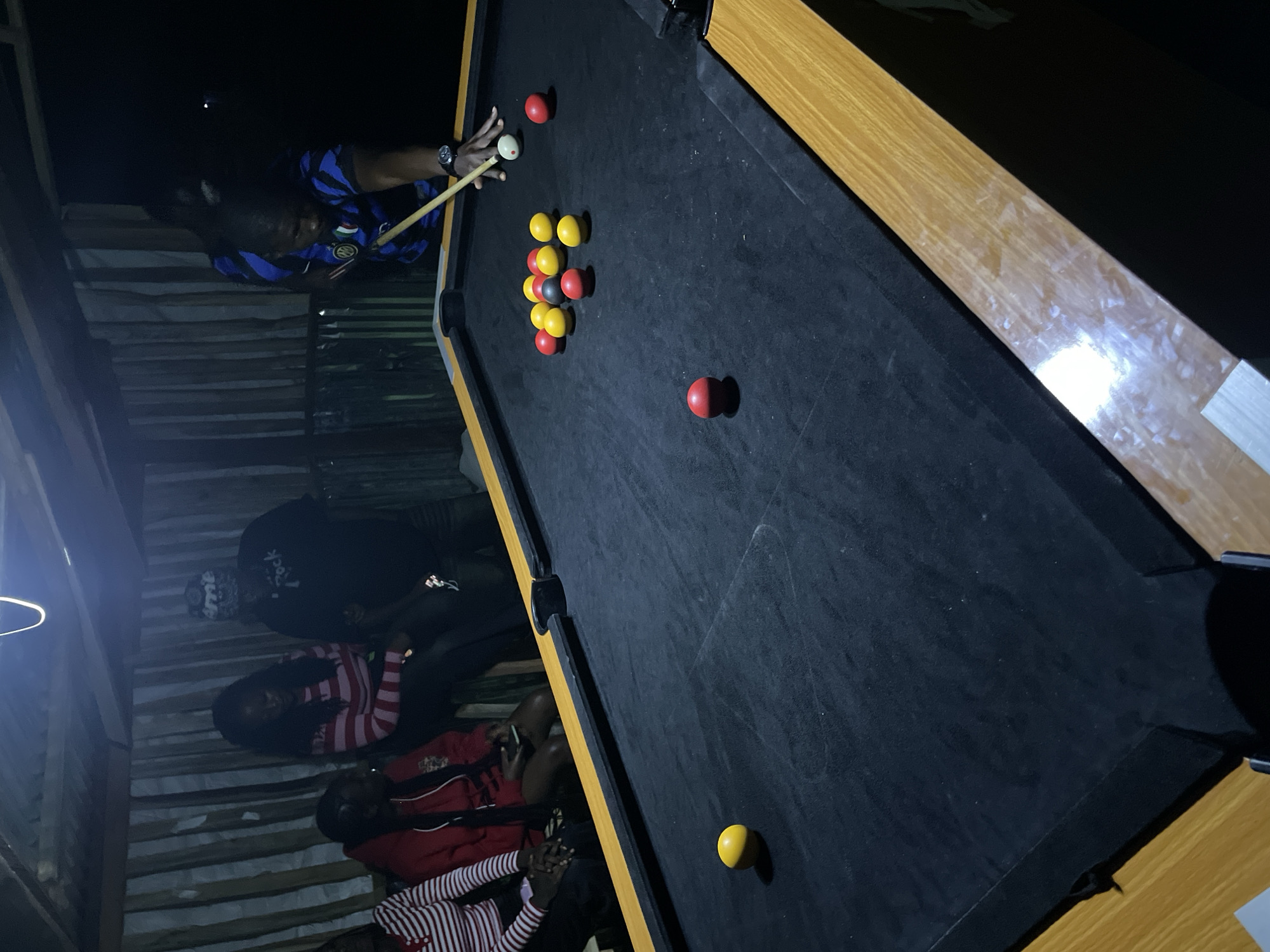 The image depicts a group of people gathered around a pool table in what appears to be an outdoor, covered area, possibly in Durumi, Nigeria. The setting is dimly lit by an overhead light fixture. Several individuals are visible in the background, seated and standing. One person, wearing a blue and black striped shirt, is actively playing pool, aiming the cue ball with a cue stick. Various colored pool balls, including red, yellow, and a black ball, are scattered on the dark green felt of the pool table. The pool table has a light brown wooden frame. The background features wooden plank walls and a corrugated metal roof, suggesting a rustic or open-air establishment.