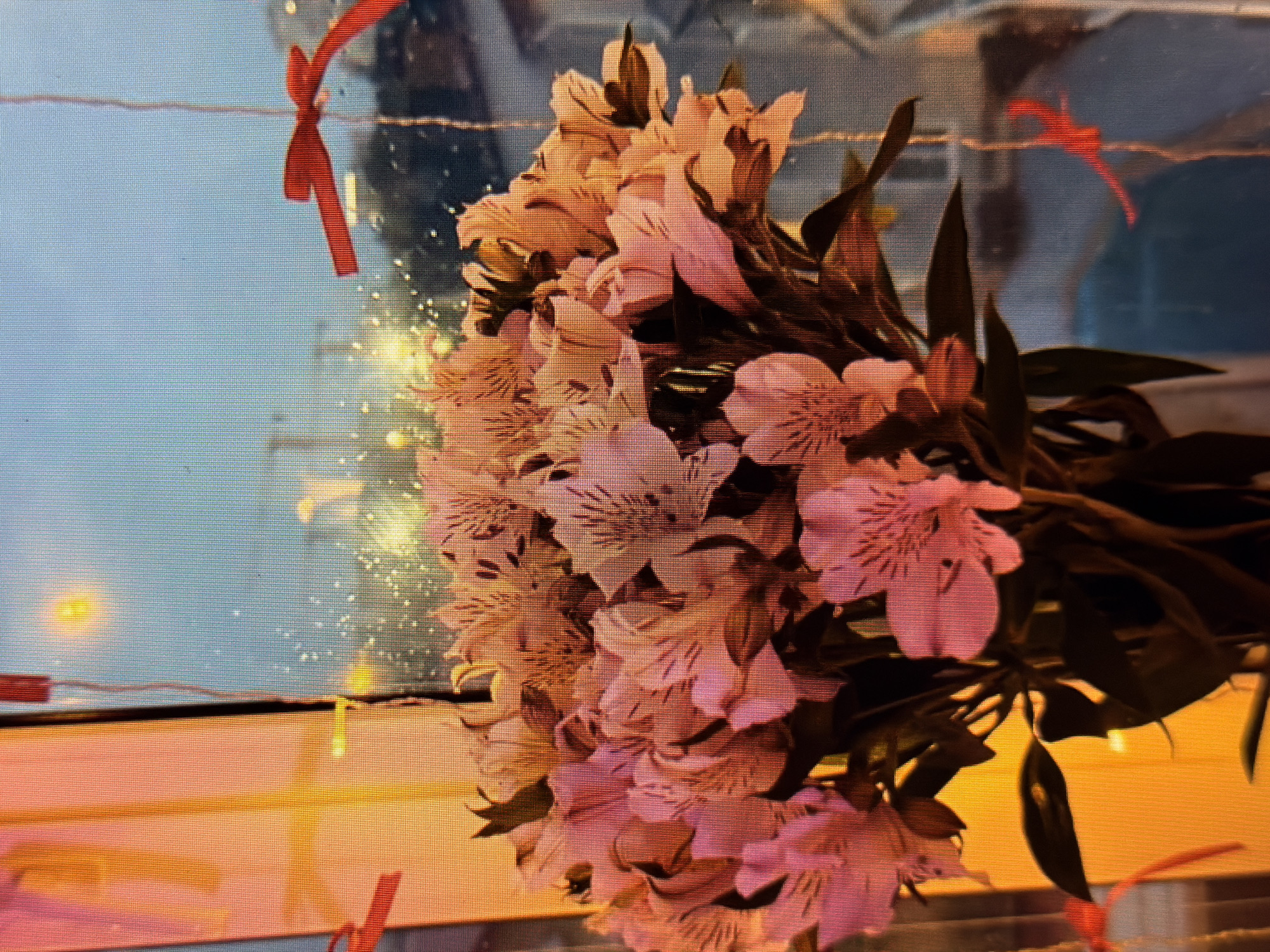 A close-up, vertically oriented, slightly blurry photograph depicts a bouquet of pale pink and white alstroemeria flowers in a vase. The flowers are the dominant subject, filling most of the frame. Red ribbons are visible, tied to what appear to be strings or wires, with one in the upper right corner and another in the lower left. The background is out of focus, showing what seems to be a window with a cityscape visible through it, featuring lights and buildings. The lighting suggests it may be dusk or evening. The location is not discernible from the image.