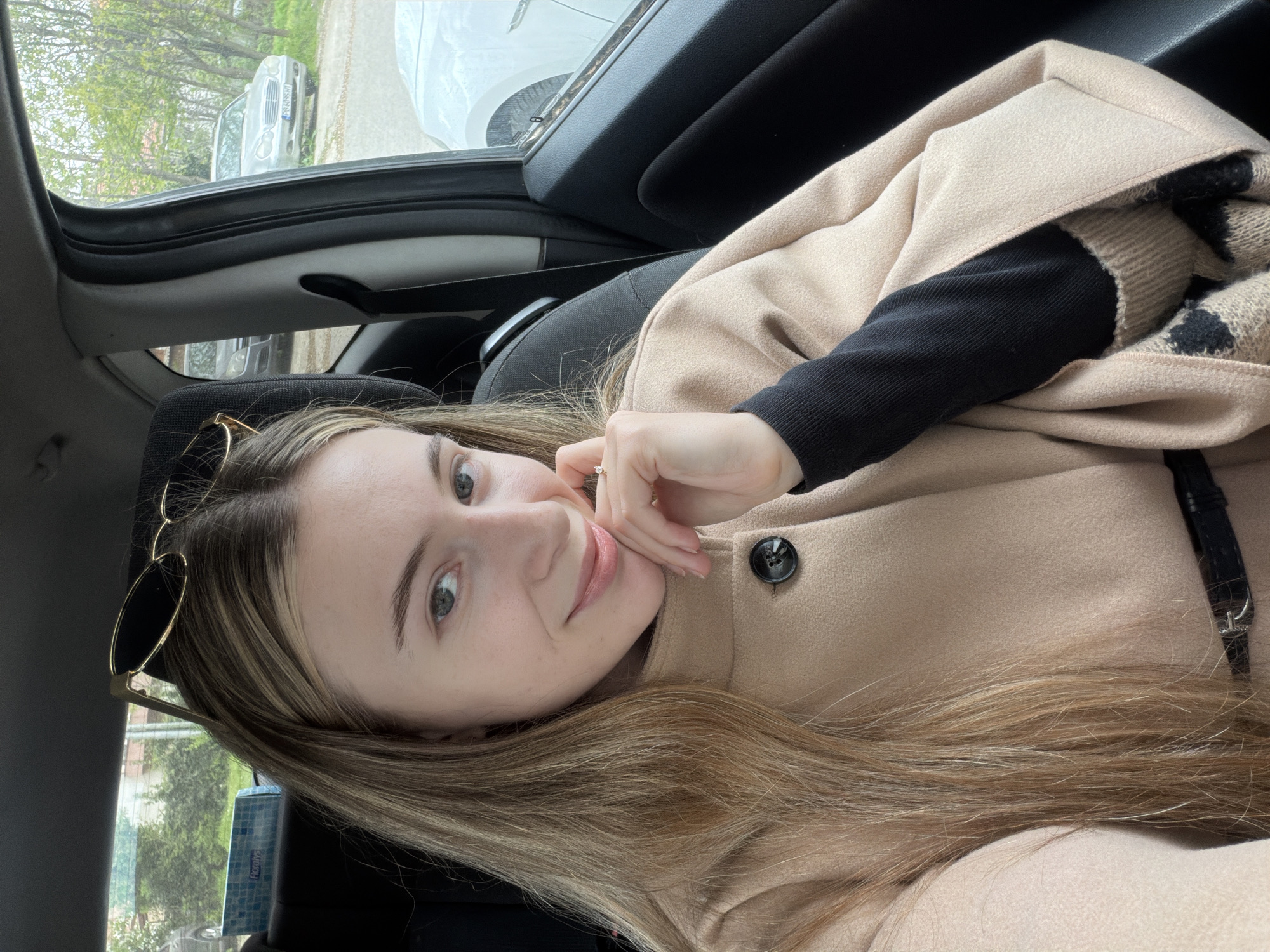 A young woman is in the driver's seat of a car. She is fair-skinned, with long blonde hair and grey eyes, and wears a beige coat over a black sweater. Sunglasses are propped on her head. Her left hand is raised to her face. The car is parked on a street in Plovdiv, Bulgaria. Another car is visible through the open sunroof. A box of Floraly's facial tissues is by the door.