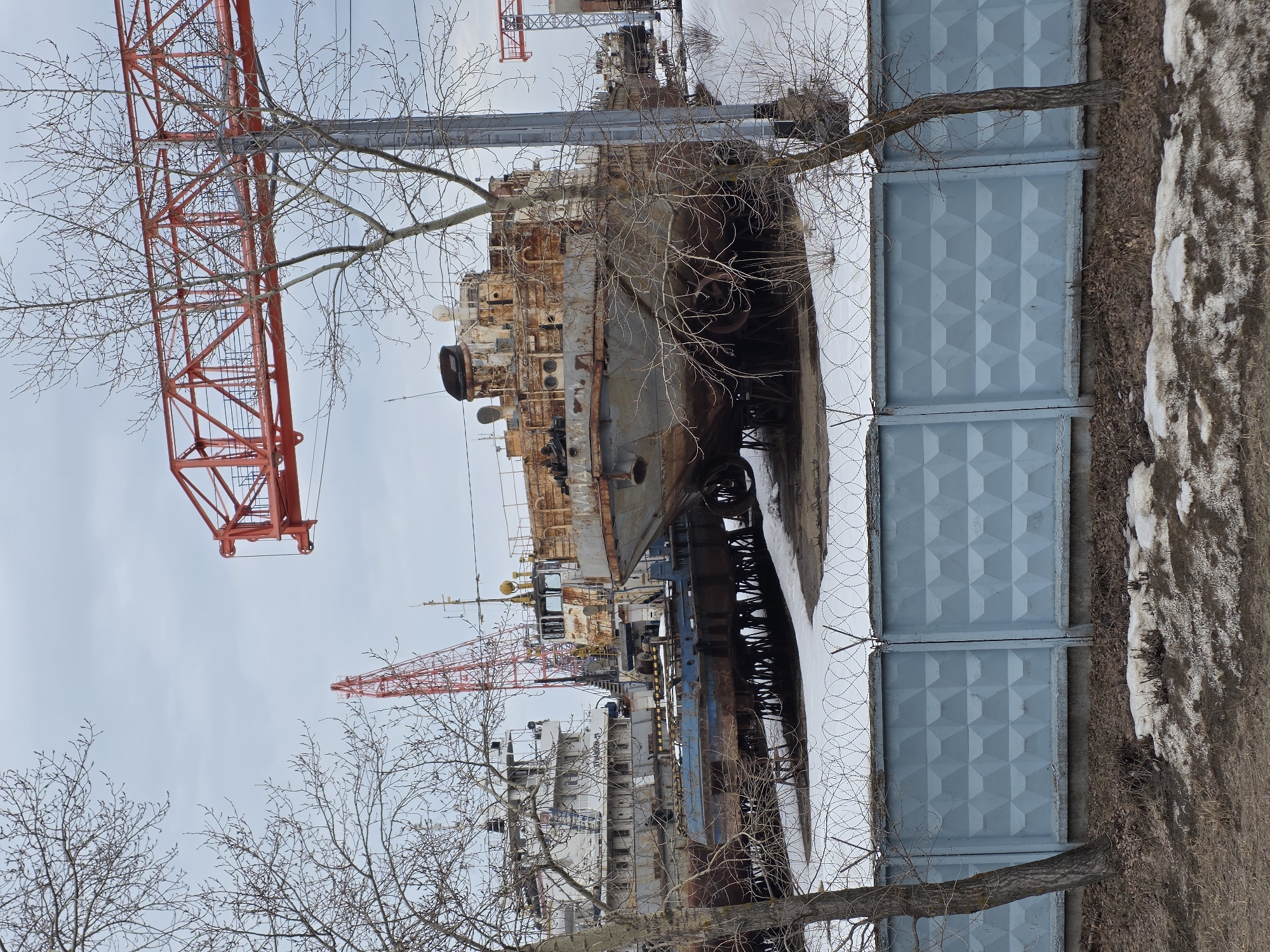 The image depicts a ship graveyard in Chaikovsky, Russia. Several rusty ships are visible, in varying states of disrepair. A large red crane stands above the ships, suggesting ongoing work or dismantling. A concrete fence runs along the right edge of the image, separating the graveyard from the surrounding area, which appears to be a mix of dirt, snow, and bare trees.