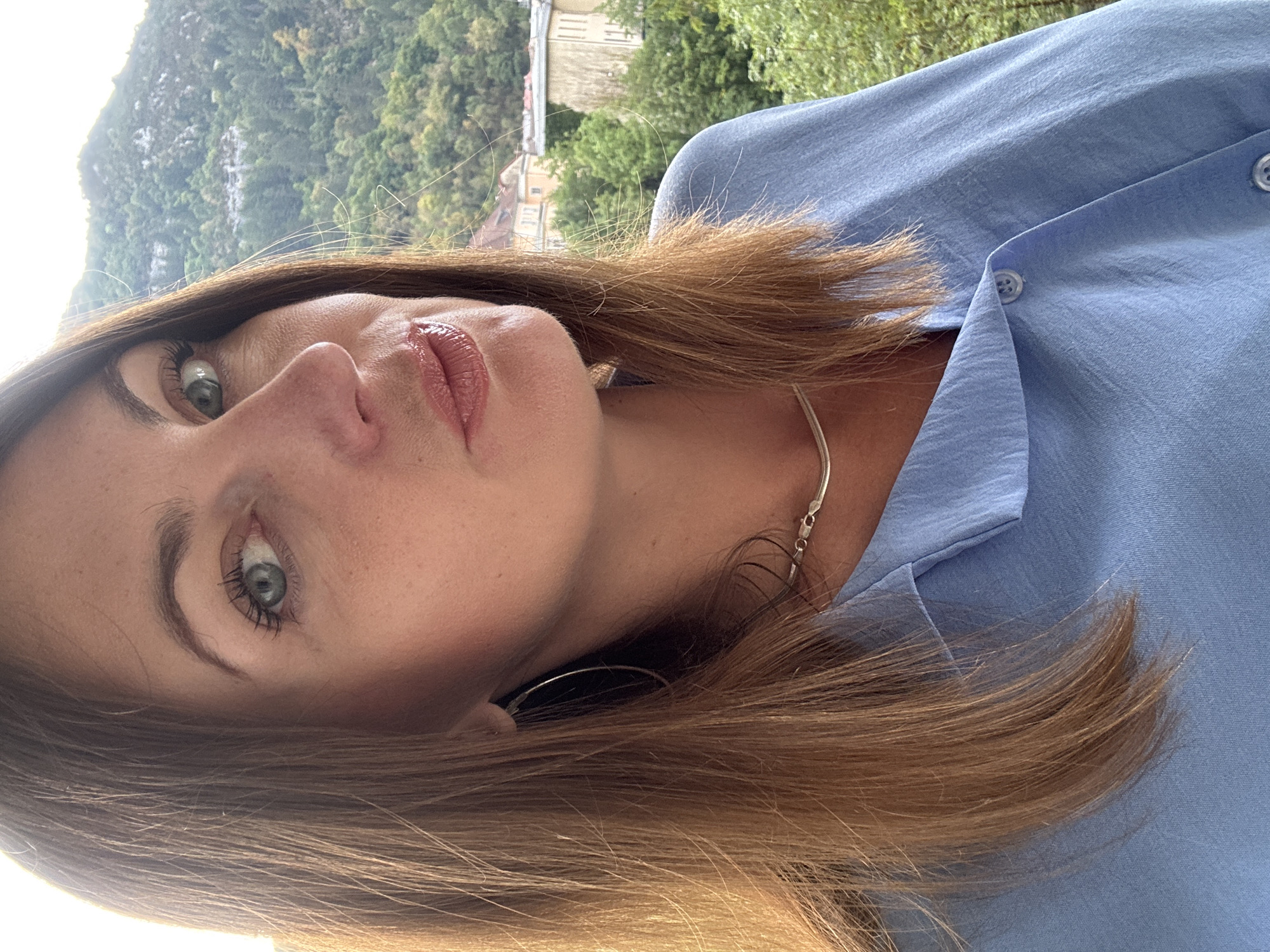 The image is a close-up, waist-up portrait of a woman taken outdoors. She has long, light brown hair and is looking towards the viewer's right. Her left eye, a light green color, is visible and appears to be wearing mascara. She has light makeup, including lip gloss. She is wearing a light blue button-down shirt and a thin silver necklace. A large silver hoop earring is visible on her left ear. The background consists of a blurry mountainous landscape with trees and some buildings, suggestive of a town nestled in a valley. The lighting indicates it is daytime, possibly late afternoon due to the angle of the light.
The specific location in Saint-Claude, France, is not discernible from the image itself. However, the background suggests a setting within the Jura Mountains, a region where Saint-Claude is located, known for its natural beauty and mountainous terrain.