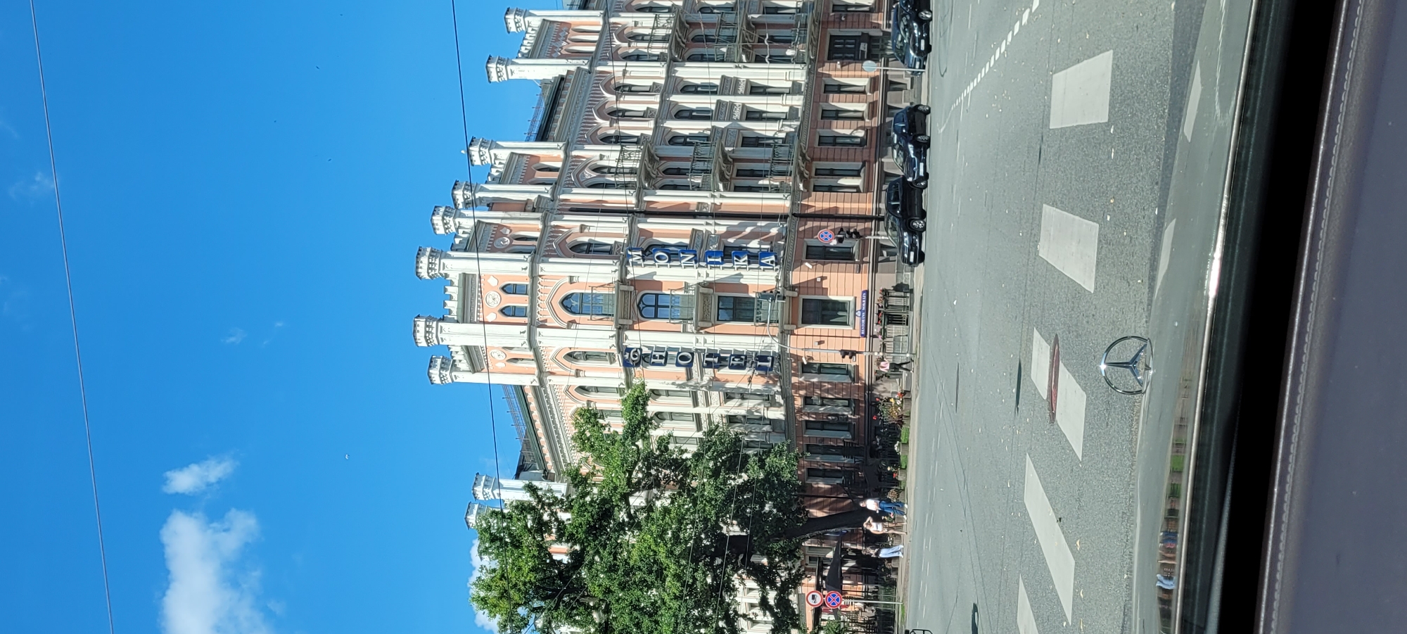 This image depicts a street view in Riga, Latvia, on a sunny day. The focal point is a grand, ornate building with pale pink and white facades, featuring gothic-inspired architectural elements like pointed arches and decorative turrets. The building is adorned with a large blue sign that reads "HOTEL MONIKA". Several cars are visible on the multi-lane street, including black sedans and a smaller black hatchback. Pedestrians are crossing a pedestrian zone in the foreground. A large green tree partially obscures the left side of the building. The sky is a clear blue with a few wispy clouds. The image is taken from a vehicle, as evidenced by the hood ornament of a Mercedes-Benz visible in the lower portion of the frame.