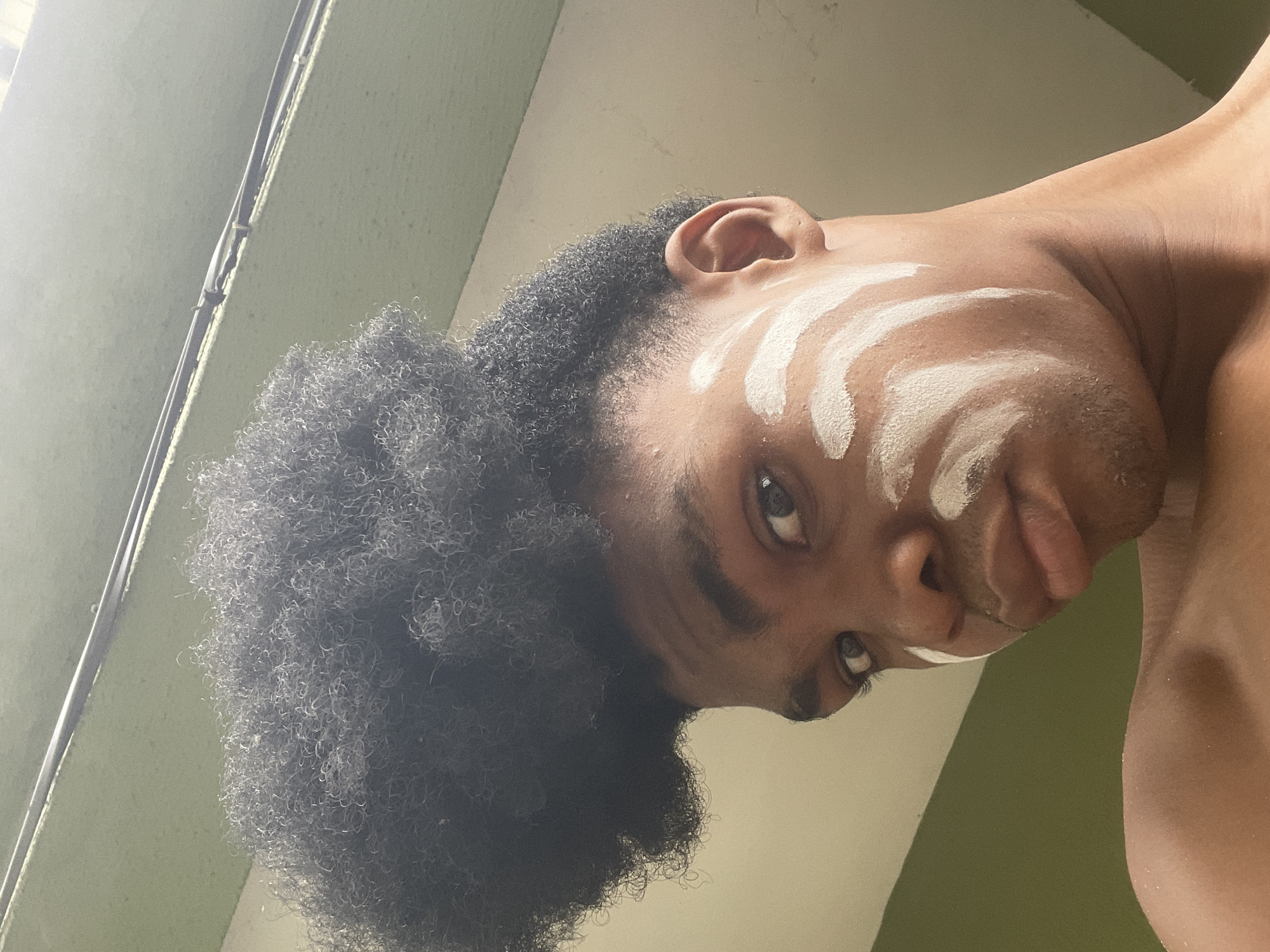A low-angle portrait of a dark-skinned person with Afro-textured hair, white paint on their face in curved lines, and a neutral expression. The background consists of a green wall, and the setting is indoors. The location is Ado, Nigeria.