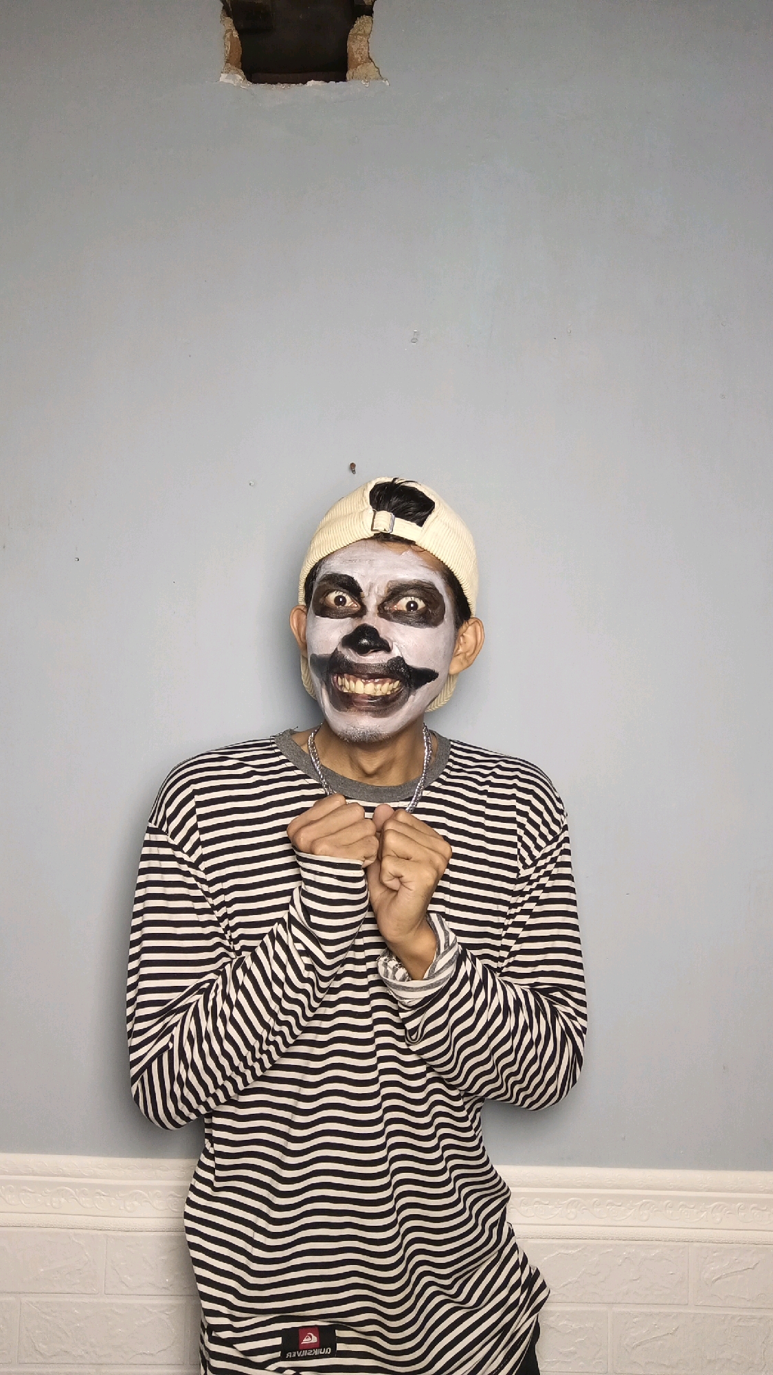 A young man wearing a skull-like face paint and a striped long-sleeved shirt stands against a light blue wall. He wears a light-colored baseball cap. His hands are clasped together in front of him. The wall has a square hole in the upper center and a decorative border near the bottom. A white brick pattern is visible at the very bottom of the frame. This image was taken in Kebumen, Indonesia.