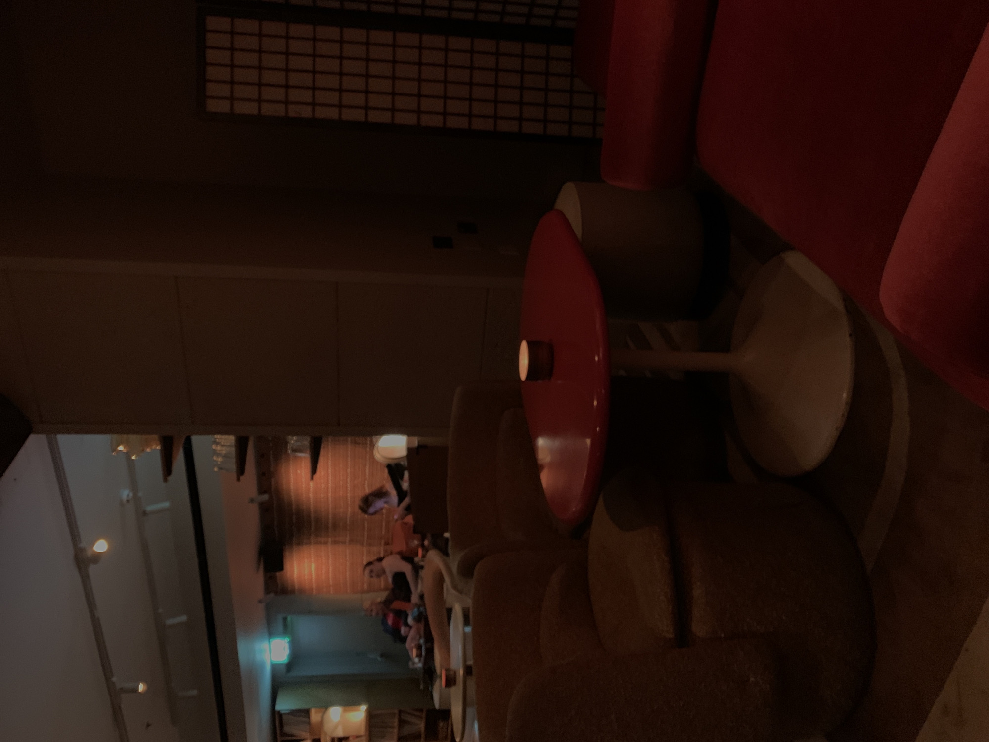 The image depicts an indoor scene in what appears to be a dimly lit, upscale bar, lounge, or restaurant during the evening. The overall atmosphere is intimate and relaxed.
In the foreground, slightly to the left, is a comfortable seating area. A plush, dark red armchair occupies the left side, with part of its back and armrest visible. Adjacent to it stands a small, round, glossy red table with a white pedestal base. A small, glowing light source, possibly a candle or a subtle lamp, is placed on the table's surface, casting a soft glow. To the right of the red table, a textured, brown armchair with a low back offers additional seating.
The background reveals a larger, more brightly illuminated area. Several people are visible here, seated at tables and engaged in social interaction, though their faces and specific activities are indistinct due to distance and the warm, low lighting. This area appears to be a more open space within the establishment, possibly a main dining or lounge section.
Architecturally, a dark wall with a distinctive grid-like pattern along its top edge spans the upper portion of the scene, adding a decorative or structural element. Various artificial light sources, including overhead lights and possibly lamps, illuminate the background area, creating a warm and inviting glow.
The scene suggests a social gathering in a sophisticated urban setting, consistent with the location context of London, United Kingdom. No discernible text is visible in the image.