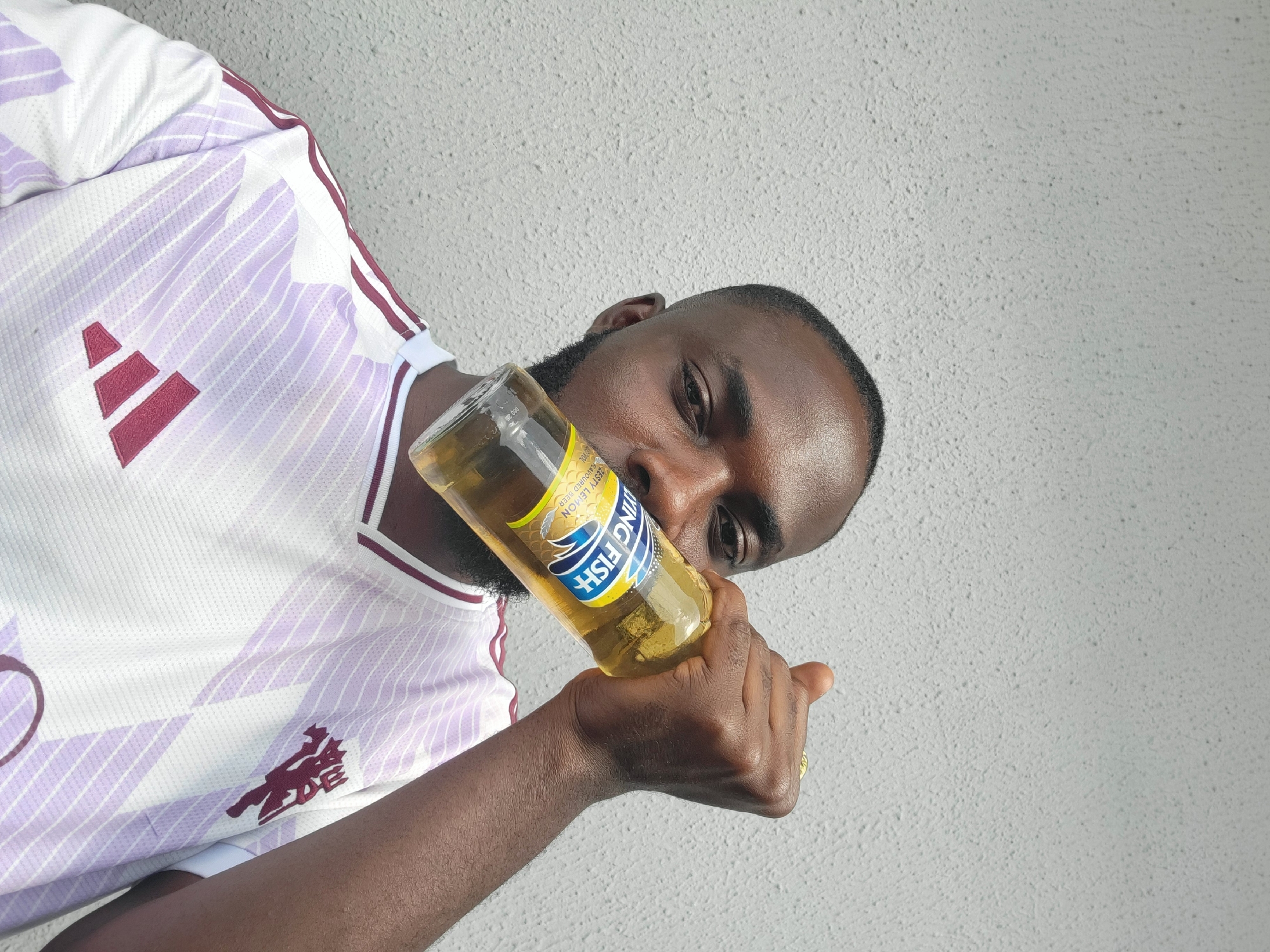 A man, likely of West African descent, is holding a bottle of Flying Fish flavored beer near his mouth, with his arm bent and hand gripping the bottle. He is wearing a white Adidas Manchester United jersey with a purple and white patterned design. The background is a textured, light gray wall, typical of urban environments in Jos, Nigeria. The lighting suggests it is daytime. The man's face is partially visible, with his left eye looking towards the camera. A ring is visible on his right hand. The overall impression is a candid or posed photograph taken outdoors against a neutral backdrop.