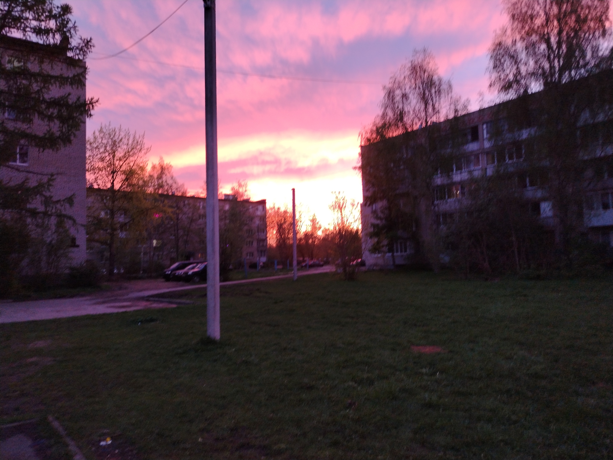 The image shows an urban landscape at dawn or dusk. The sky is a vibrant mix of pink and purple hues, suggesting a sunrise or sunset. Two multi-story buildings flank the scene, one on each side. A grassy area occupies the foreground, and a concrete path runs along the left side, with several parked cars visible. A utility pole stands prominently in the center of the frame.