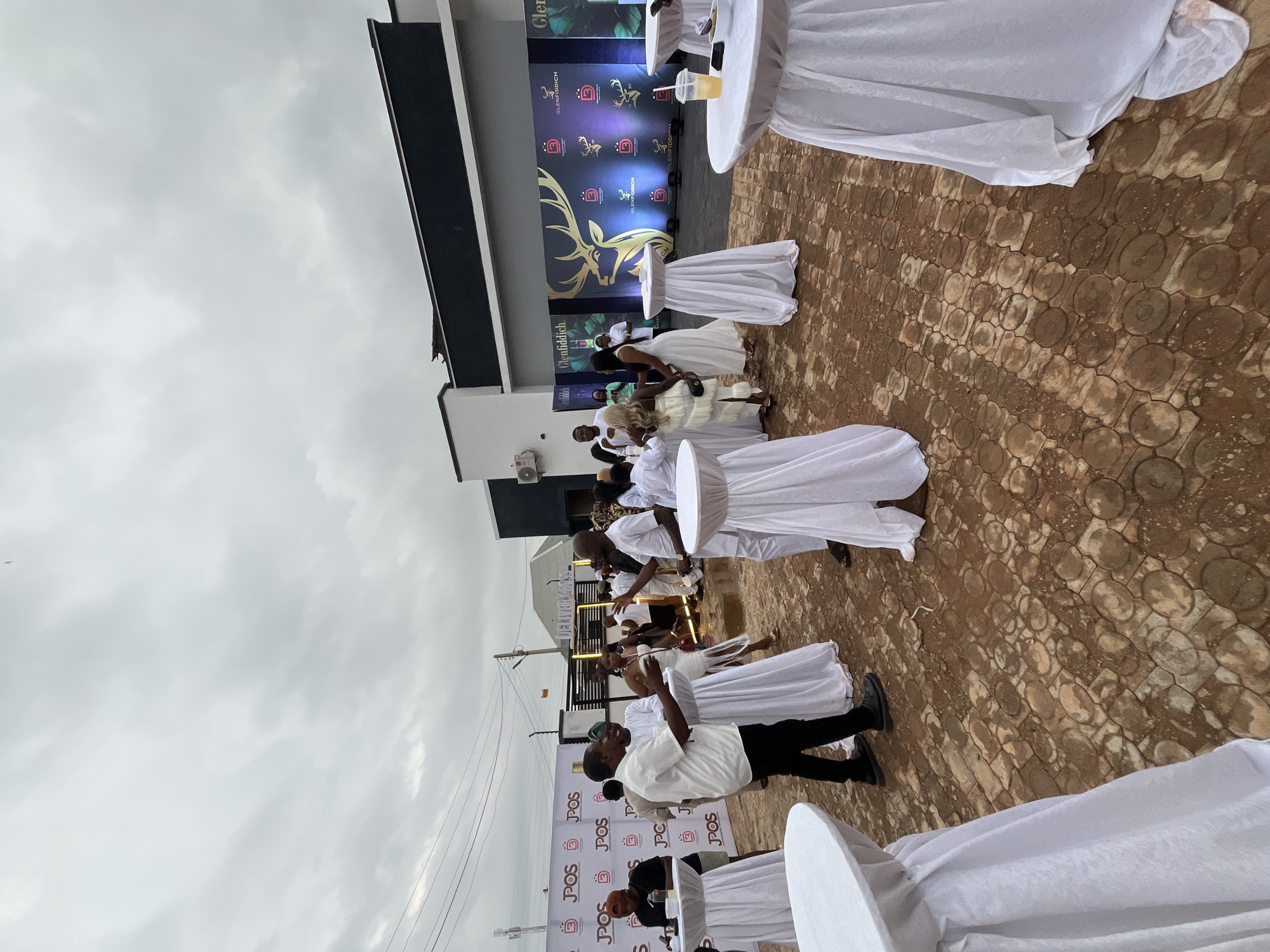 The image depicts an outdoor event at a venue, possibly in Bar Gada, Nigeria, characterized by a large, modern building with a dark facade and white accents. A banner displaying "Glenfiddich" with a stag logo and "JPOS" branding is prominently featured. Guests, dressed in predominantly white attire, mingle and converse around high-top tables draped in white cloths. Some individuals are captured in mid-conversation, while others appear to be observing the surroundings or engaging with drinks. The ground is paved with a patterned brick or stone material. The sky is overcast, suggesting a cloudy day. Essential details about the location include the presence of a building with a corporate or event-space aesthetic, outdoor seating arrangements, and branding indicative of a sponsored event. The atmosphere appears social and celebratory.