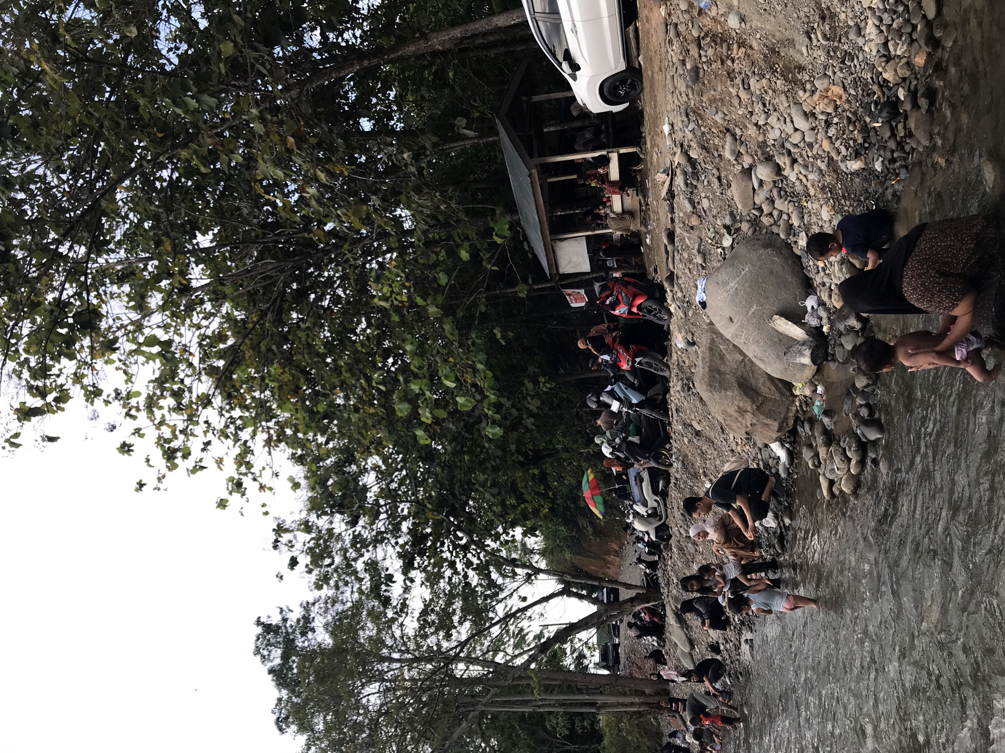 The image depicts a river scene near Meurah, Indonesia. A white car is parked near a small, open-sided structure. Several motorcycles are parked nearby. People are gathered along the riverbank and in the water, with some appearing to be washing or playing in the river. The riverbank is rocky, and trees provide shade.