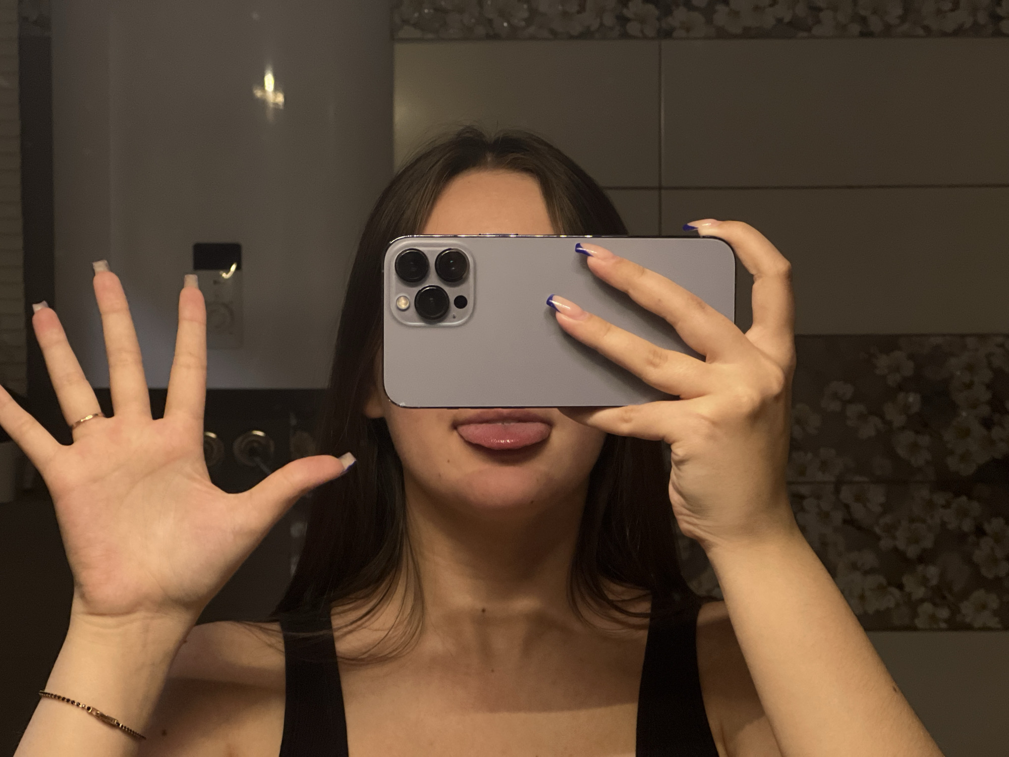 A young person, likely female, is captured in a mirror selfie, obscuring most of their face with a grey/silver iPhone, which appears to be a Pro Max model. Their long, dark hair frames the visible lower part of their face, where their tongue is playfully sticking out. They are wearing a black tank top or camisole.
The smartphone is held horizontally with the right hand, featuring a distinct camera module with three lenses and a flash visible on its back. The fingernails on this hand are painted with blue tips in a French manicure style. The left hand is raised towards the camera, fingers splayed, displaying a simple ring on the fourth finger and a dark, possibly beaded, bracelet on the wrist. The nails on the left hand appear natural or have a clear/light polish.
The background, reflected in the mirror, consists of dark-grey tiled walls. Some sections feature decorative tiles with a subtle floral or abstract pattern in lighter tones. A dark rectangular object, possibly an electrical outlet or switch, is visible in the reflection behind the raised left hand, along with a bright light reflection. The lighting suggests an indoor setting. The overall mood is casual and playful. The location context is Chaikovsky, Russia.