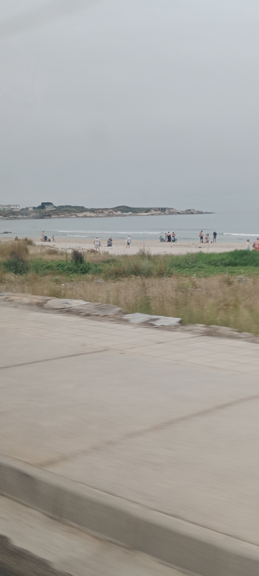 The image captures a wide shot of a beach scene on an overcast day, likely in Ribadeo, Spain. In the foreground, a light-colored paved surface, possibly a walkway or road, occupies the bottom half of the frame, showing motion blur. Beyond this, a strip of dry, yellowish grass and some green vegetation borders the sandy beach. The beach itself is populated by several individuals and small groups. Some people are standing, while others are seated. Further out, the sea is visible with gentle waves breaking on the shore. In the distance, across the water, a rocky coastline with vegetation and a few buildings can be seen. The sky is a uniform, pale grey, indicating cloudy weather. The overall impression is a relaxed, casual day at the beach with limited activity.