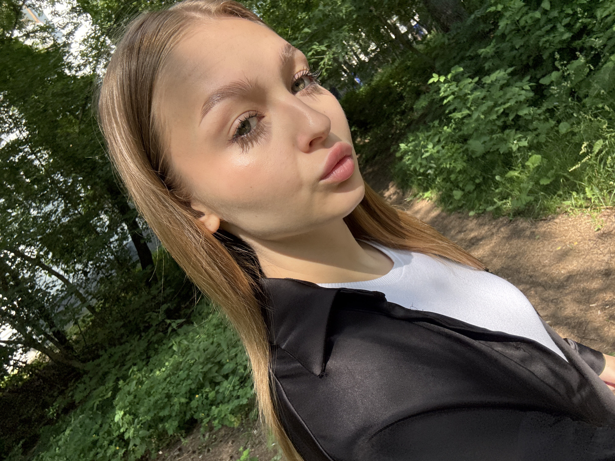 This is a selfie taken by a young woman with long, blonde hair. She is wearing a white t-shirt and a black open shirt. Her face is turned to the side, and she is looking towards the camera with a playful expression, puckering her lips. The background is a lush green forest with dappled sunlight filtering through the trees, suggesting it is daytime and the weather is pleasant. The setting appears to be a park or woodland area, with a dirt path visible on the right side of the image. The light suggests it could be late afternoon. There is no visible text in the image.