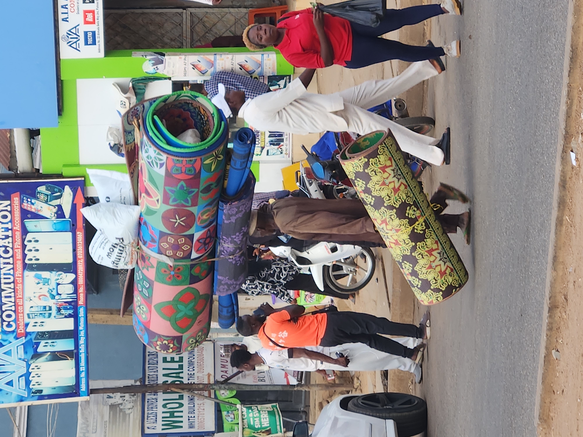 This media file captures a lively street scene in Jos, Plateau State, Nigeria, during the daytime. The setting is a commercial area with shops lining a paved road and dirt shoulder.
Several people are present, indicating everyday activity. A woman in a red top and dark trousers walks away from the camera, carrying a black bag. Near her, a man dressed in white attire, including a white cap, also walks along the street. Further down, another man in an orange t-shirt and dark pants is standing or moving.
A dominant feature of the scene is a collection of colorful, vibrantly patterned rolled-up mats or rugs, possibly for sale or transport. They are seen leaning against buildings and standing upright on the sidewalk. A white motorcycle is parked near the street. The foreground shows a car tire, suggesting a parked vehicle or general street elements.
The background is filled with various signs for businesses. A prominent blue and green sign for "AVA COMMUNICATION" advertises "Dealers on all kinds of Phone and Phone Accessories," explicitly stating its address as "No. 22 Ahmadu Bello Way Jes, Plateau State," along with phone numbers (08064728972, 07036124867). The sign also displays phone brands like "TECNO" and "itel." Another sign indicates "AS ALEXIS PHONES, A CORIES AND REPAIRS" and "WHOLESALE" with additional contact details. Other visible text includes "REDMI A7 Pro," "SIM CARD & SIM SWAP HERE," and "EASY BUY." The architecture includes buildings with sections painted in bright green and blue.
