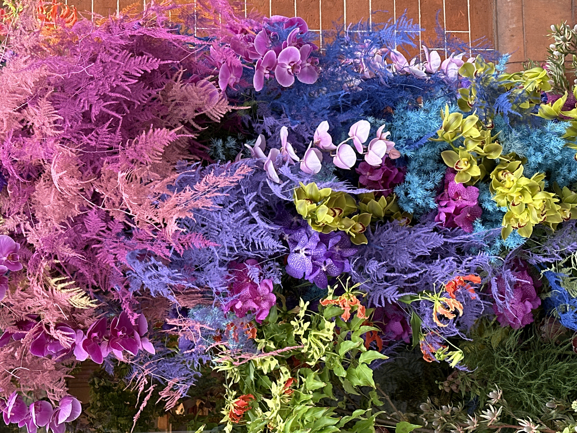 The image shows a close-up of a floral installation. The background consists of a brick wall with a white wire grid attached to it. A variety of flowers and greenery are arranged within the grid. Colors include shades of pink, purple, blue, green, and yellow. Specific flowers present are orchids and flame lilies. The location appears to be in London, United Kingdom, based on the style of the floral design which is reminiscent of displays found in that city.