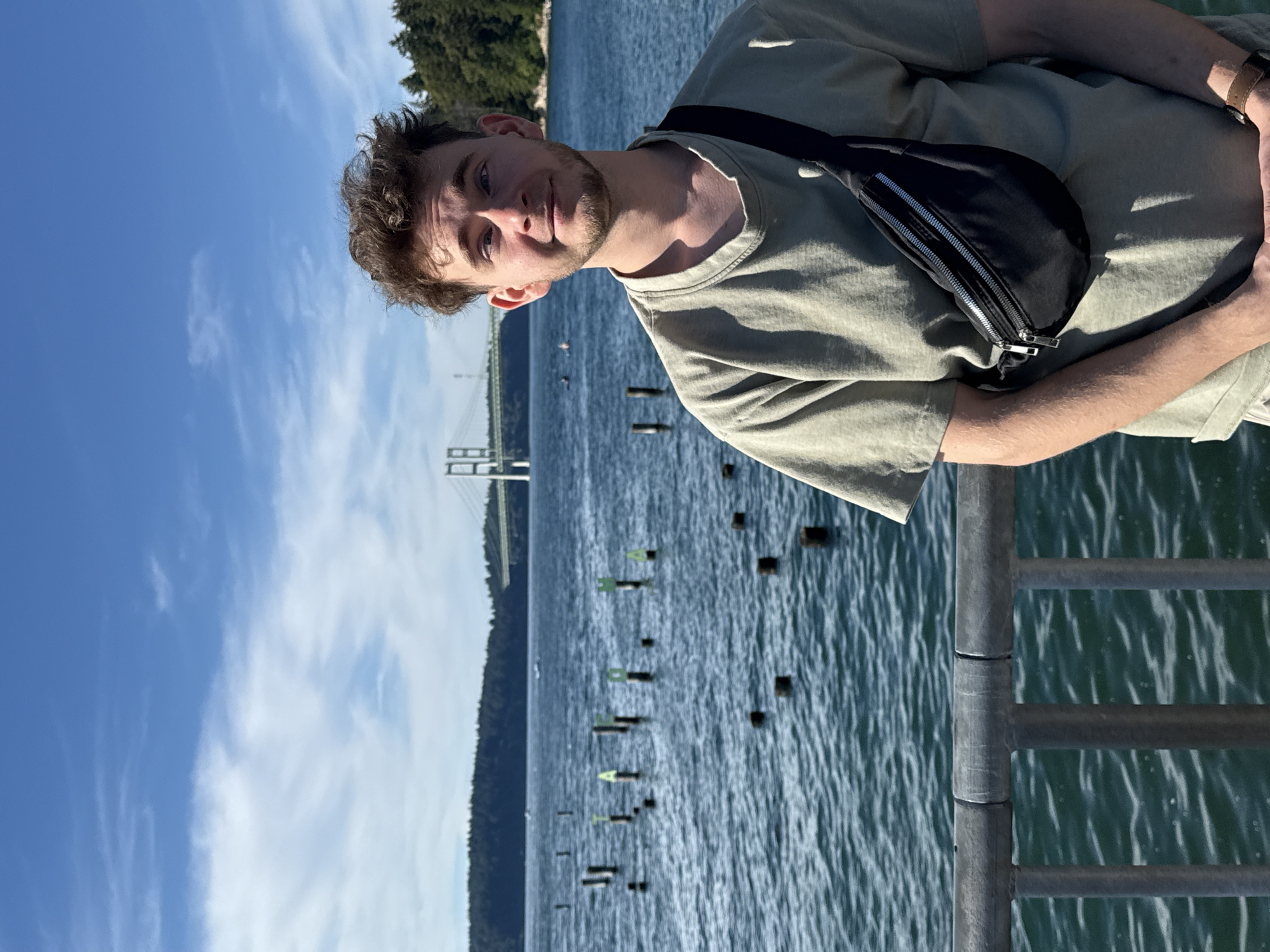 A young man with curly brown hair and a light beard is standing on a pier overlooking a body of water. He is wearing a light olive green t-shirt and a black fanny pack across his chest. The water has several wooden pilings sticking out of it, with some appearing to have green letters on them that spell "TACOMA". In the background, a large suspension bridge is visible, along with a tree-covered shoreline. The sky is blue with scattered white clouds. The overall scene suggests an outdoor recreational setting in Tacoma, Washington, USA.